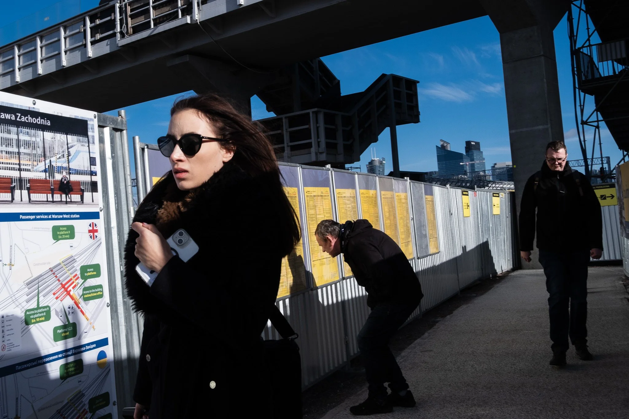 People at a train station with construction fencing and city skyscrapers in the background.