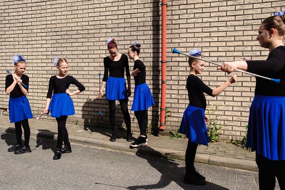 Group of young girls in black tops and blue skirts practicing twirling or dance moves outdoors against a brick wall.