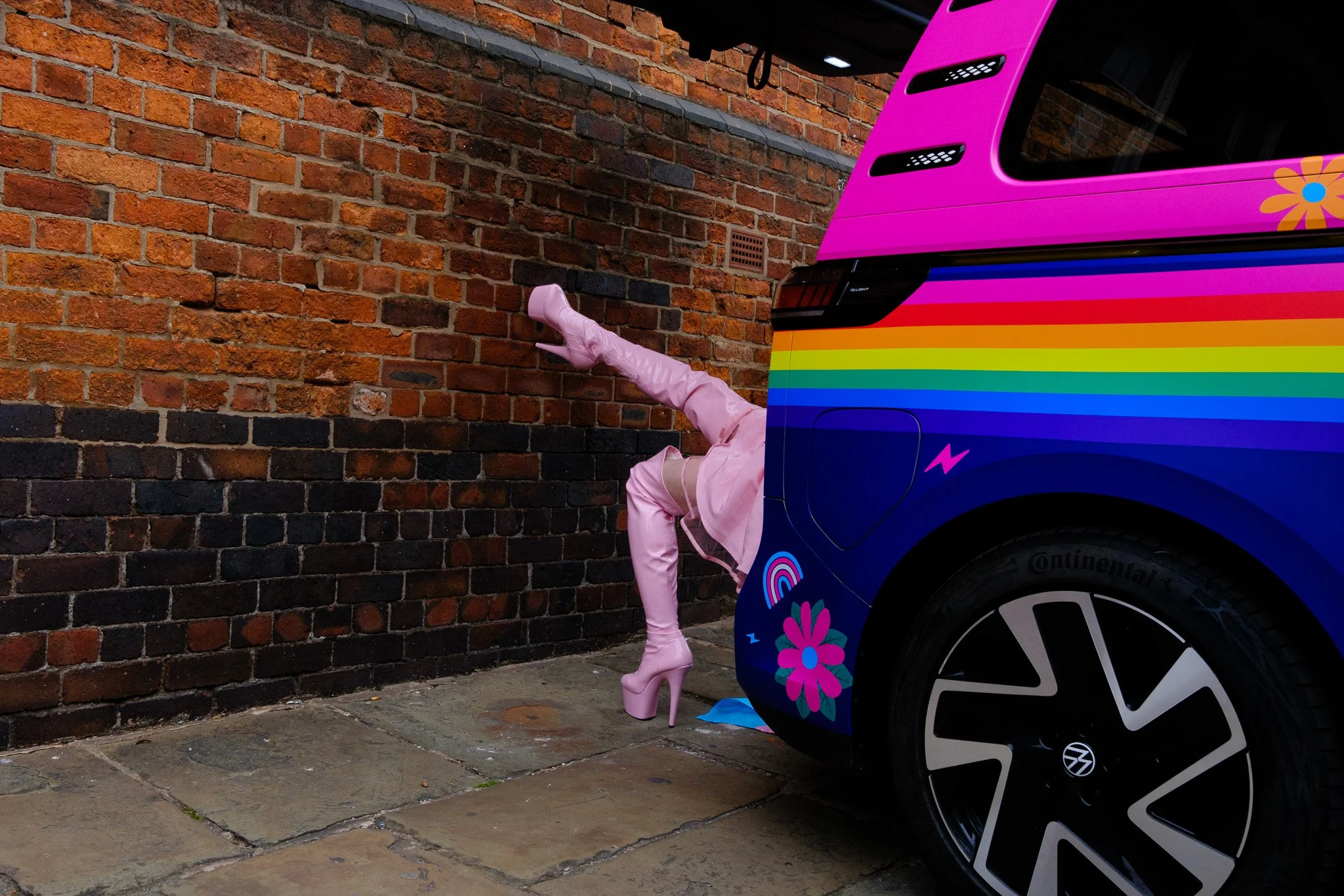 A person's leg sticking out from behind a colorful van with rainbow stripes and floral designs. The person is wearing pink thigh-high boots and pink clothing, and is hiding behind the van, which is parked against a brick wall.