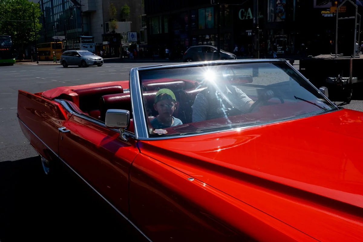 A red convertible car with two people inside, a child and an adult, on a city street with traffic and buildings in the background.