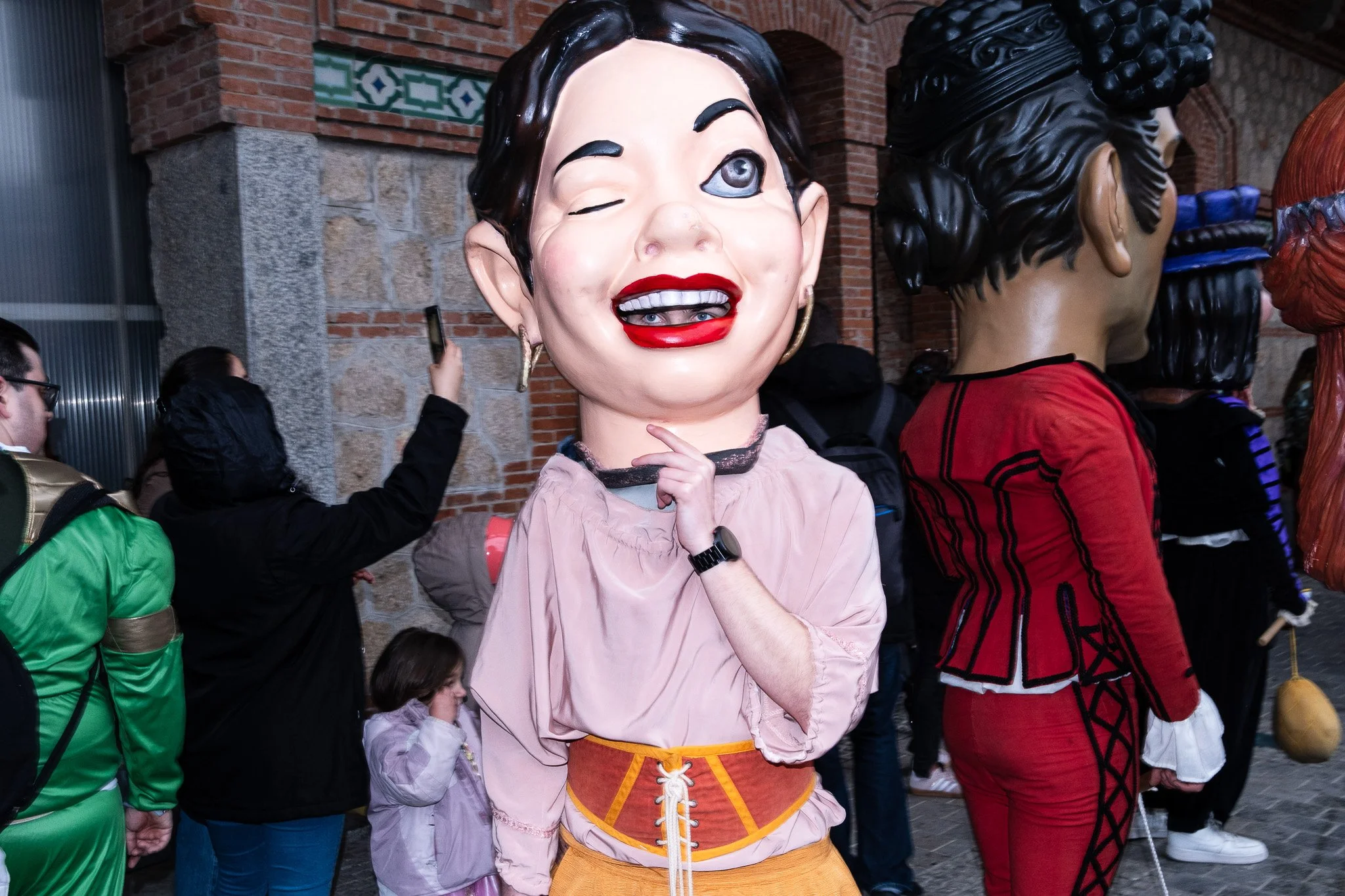 People wearing large caricature masks at a festival, with a woman in a yellow skirt and pink blouse in the center.