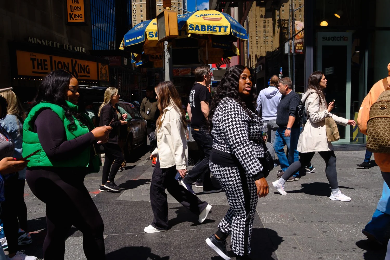 Crowd of pedestrians crossing the street in an urban downtown setting on a sunny day, with storefronts, signs, and tall buildings surrounding them.