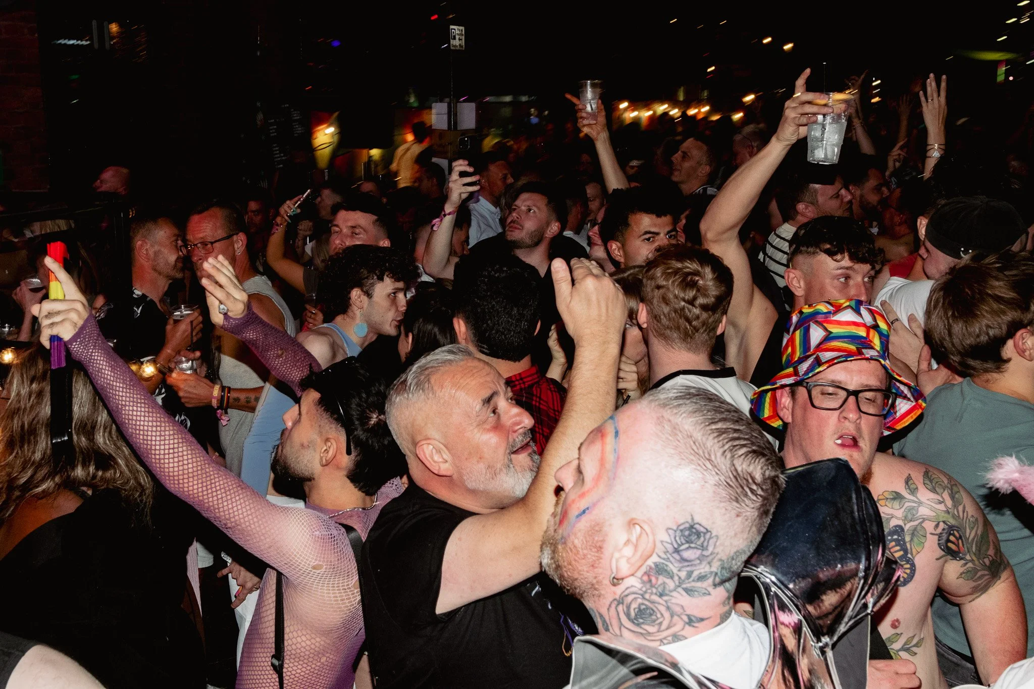 A crowded party scene with people dancing and celebrating; some are holding drinks, and one person is wearing a colorful hat