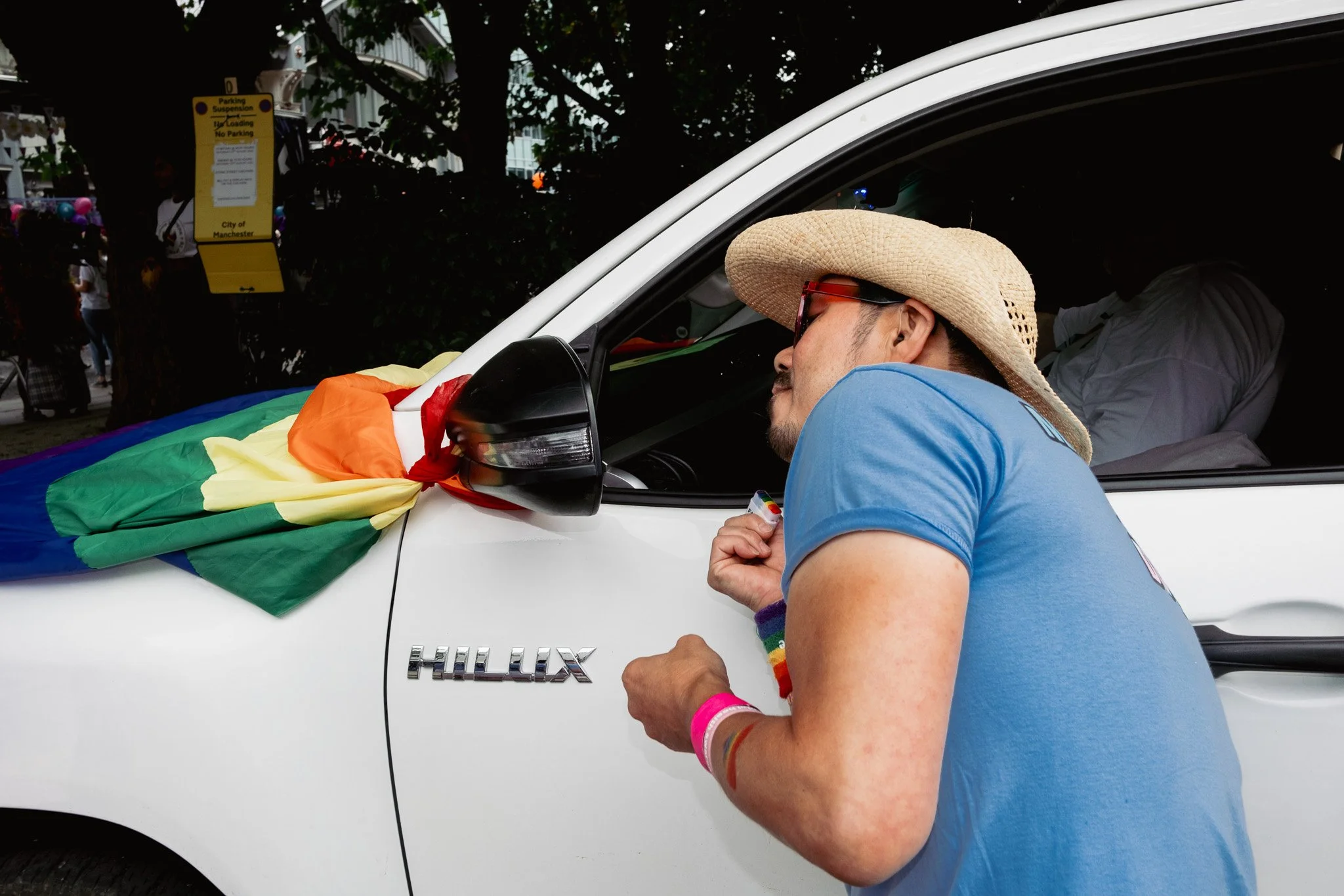 Person wearing a straw hat, sunglasses, and a blue T-shirt leaning toward a decorated white car at a pride event.