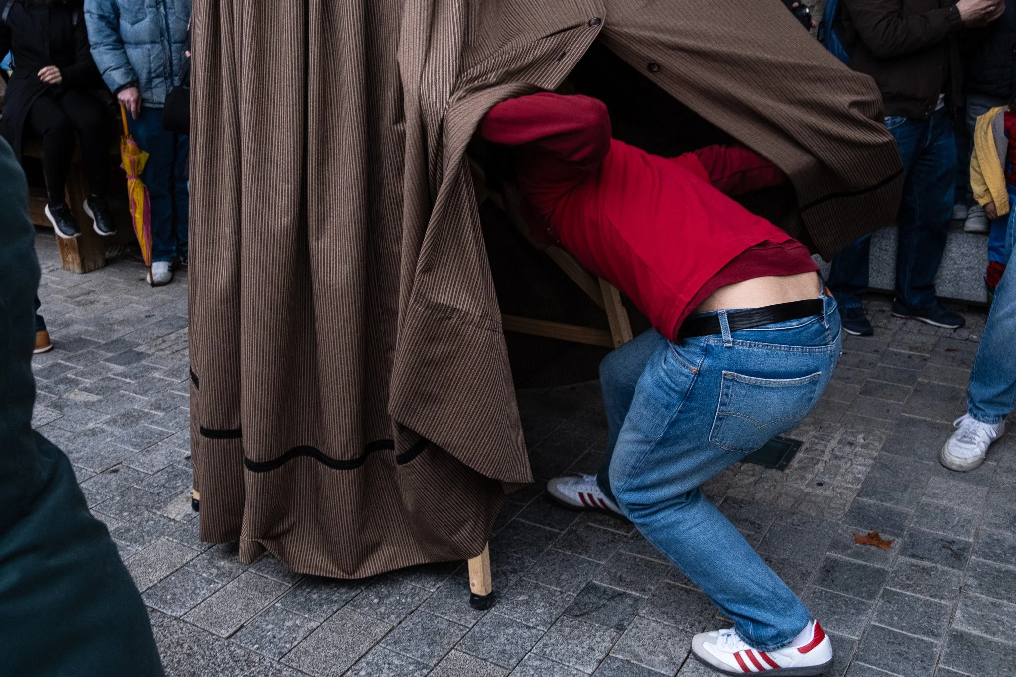 A person with a red hoodie and blue jeans appears to be under a large draped brown cloth, possibly part of an event or performance, with onlookers surrounding them.