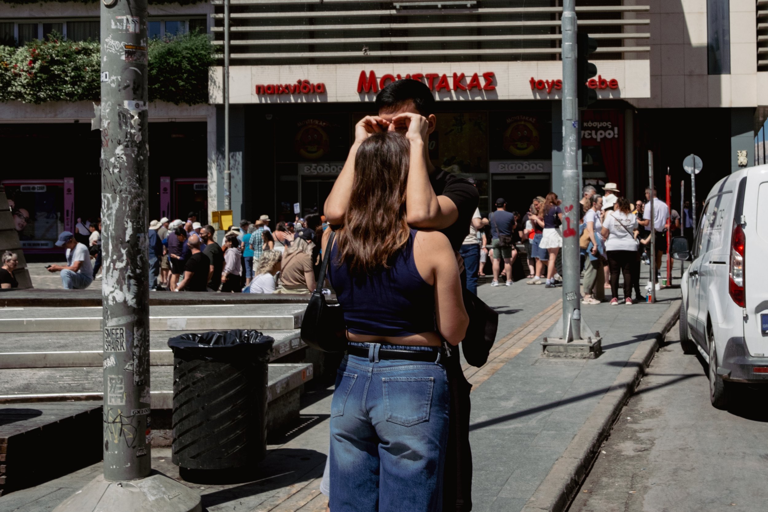 Couple hugging and kissing on a busy city sidewalk in front of a building with a toy store entrance; many people line up outside.