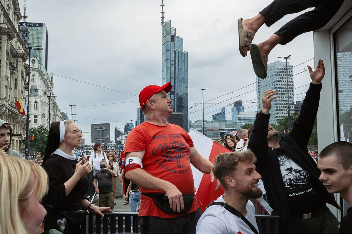 Crowd of people gathered outdoors in an urban area with tall buildings in the background. A man in a red cap and red shirt stands among them, and a person is throwing flip-flops from above. Various people are observing and participating, some with fl