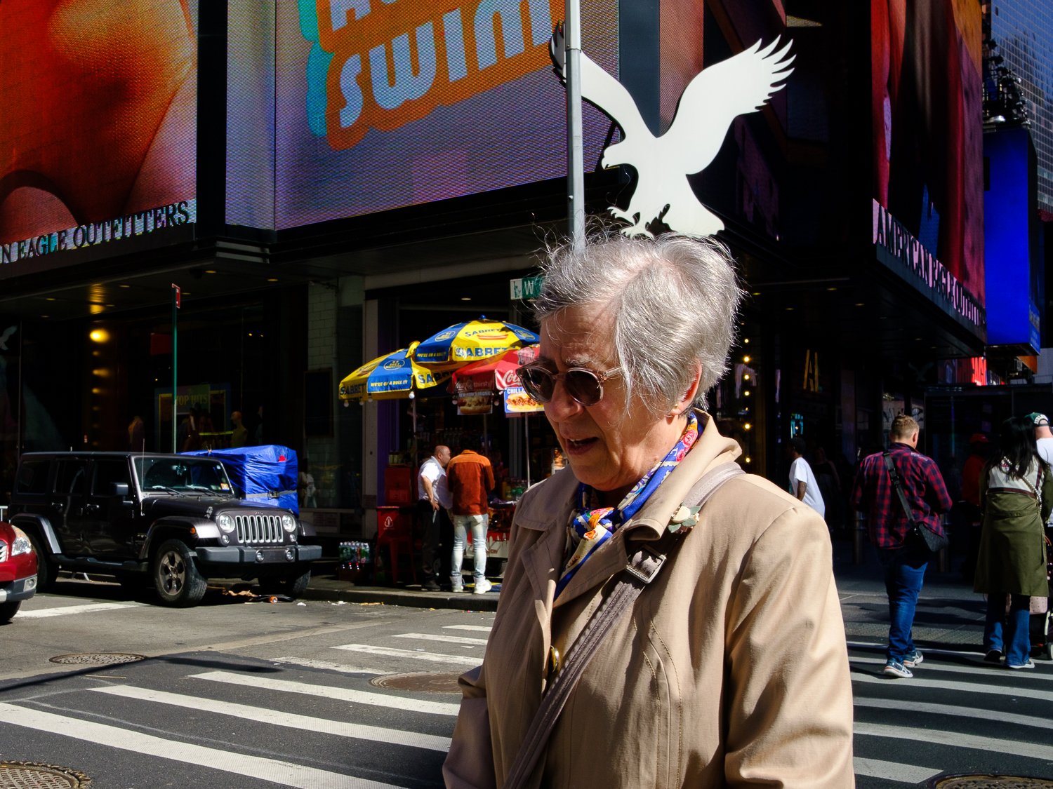 An elderly woman with white hair, wearing sunglasses and a beige jacket, walking across a busy city intersection with digital billboards in the background.