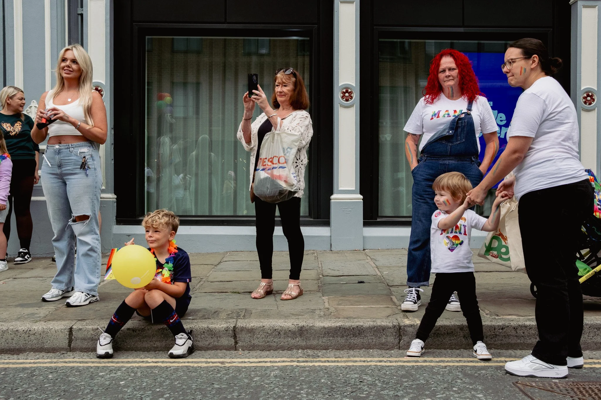 Group of diverse people, including children and adults, celebrating on a city sidewalk decorated with rainbow-themed accessories, with an event or parade taking place.