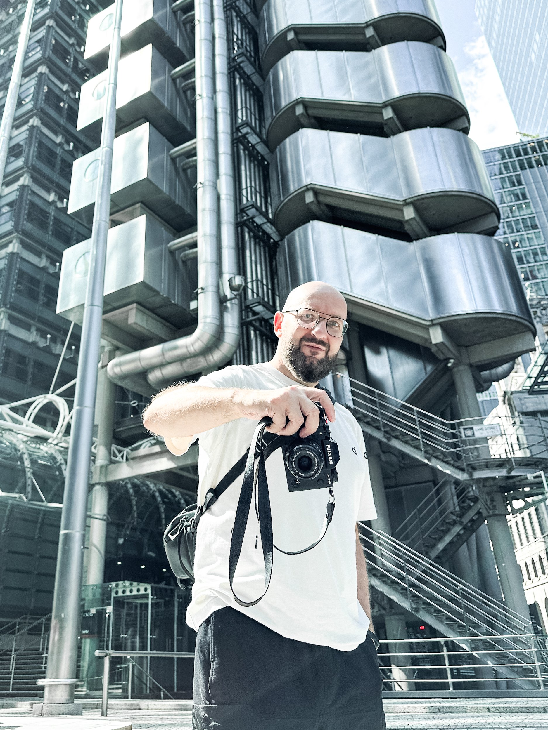 A man with a camera hanging around his neck stands in front of a modern building with rounded, metallic balconies and industrial pipes in an urban setting.
