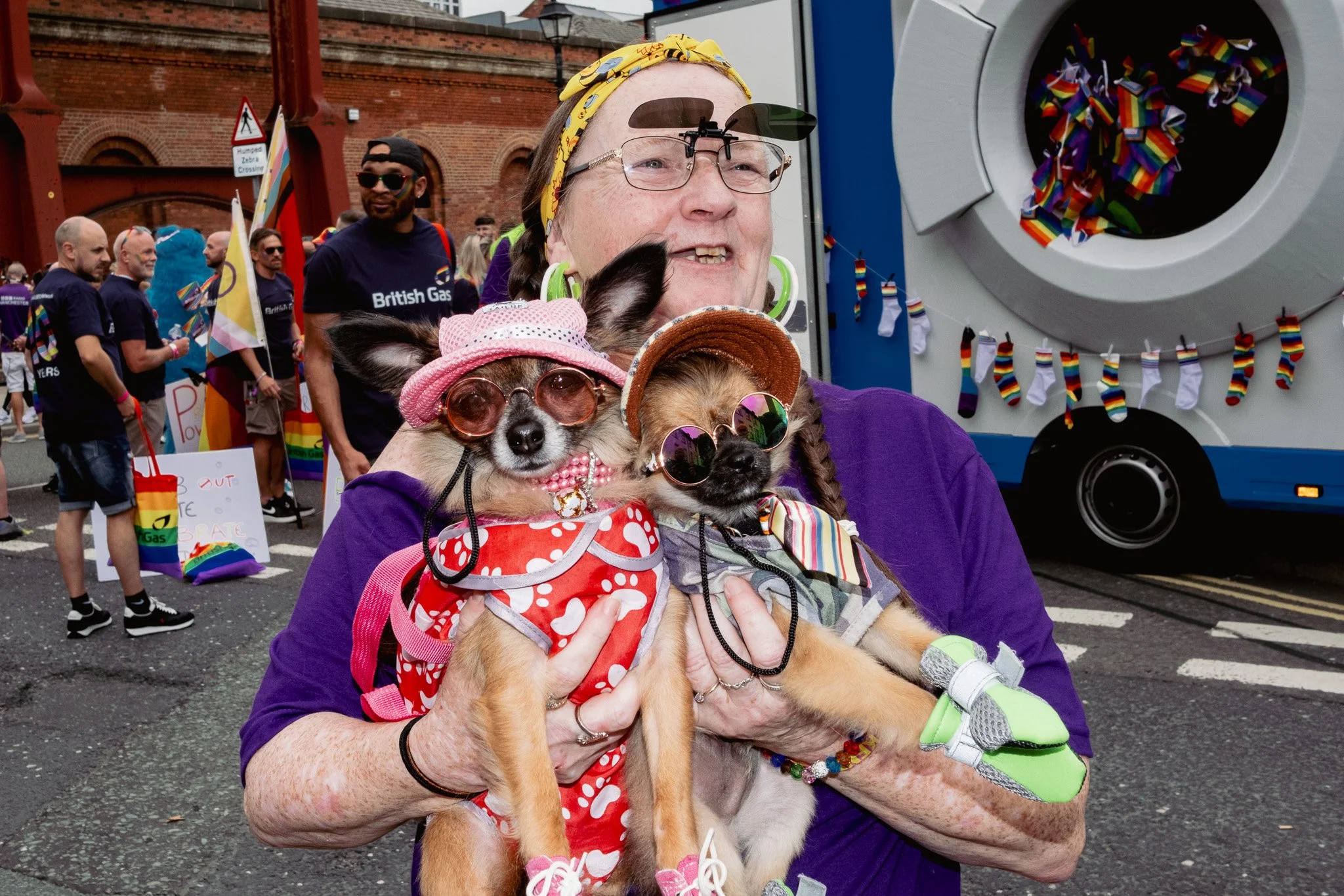A woman holding two dogs dressed in colorful costumes at a pride parade. The woman is wearing glasses, a yellow headband, and a purple shirt. The dogs are wearing sunglasses, hats, and rainbow-themed accessories. In the background, there are people w