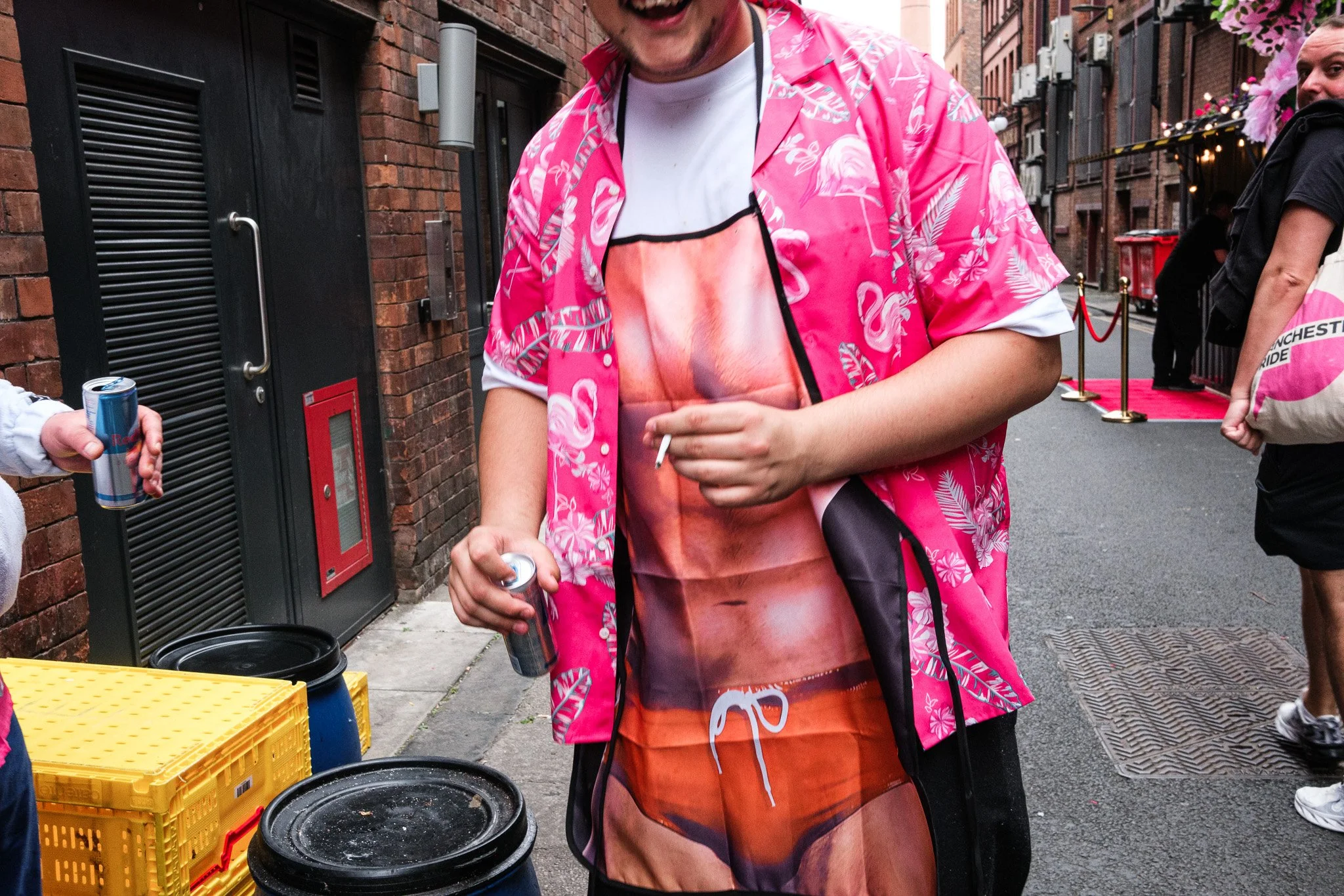 A man wearing a vibrant pink Hawaiian shirt and a swimsuit featuring a sunset and ocean scene, standing on a city sidewalk, holding a can of drink in each hand, with other people and urban buildings in the background.