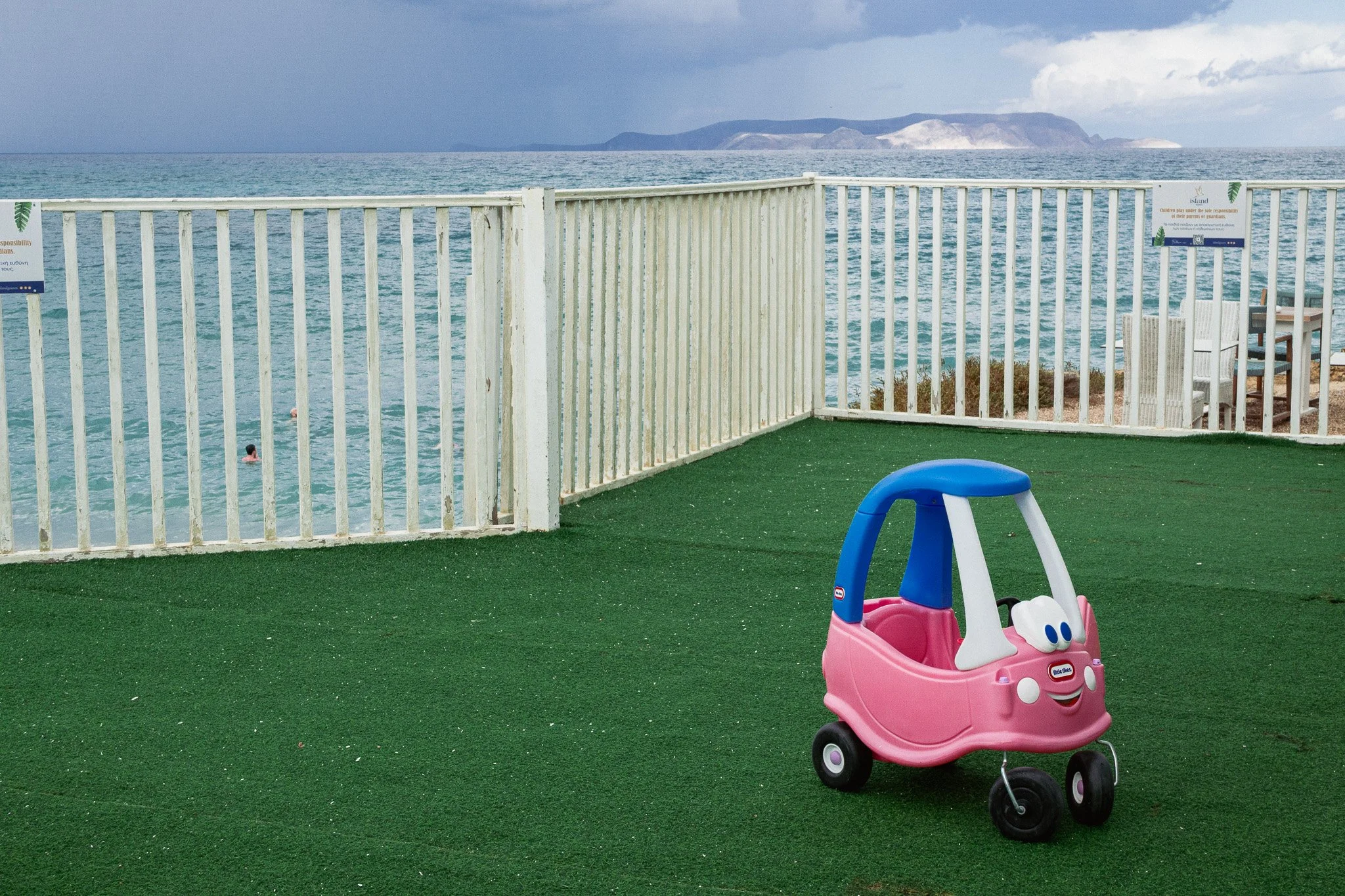 Empty pink and blue children's ride-on toy car on green turf near a white fence with an ocean and island in the background.