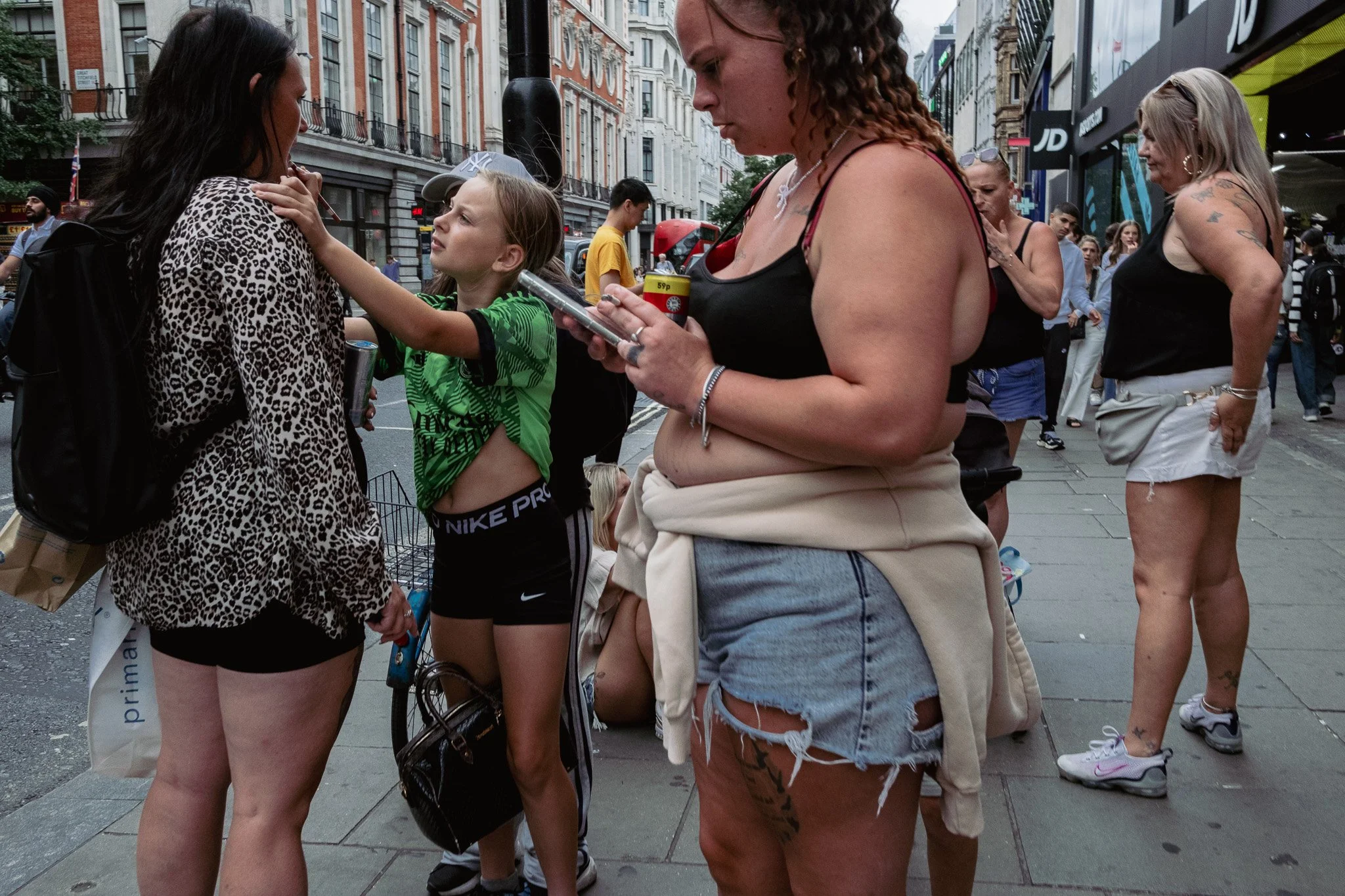 A group of women and a girl on a busy city street, some engaging in conversation, with building storefronts and pedestrians in the background.