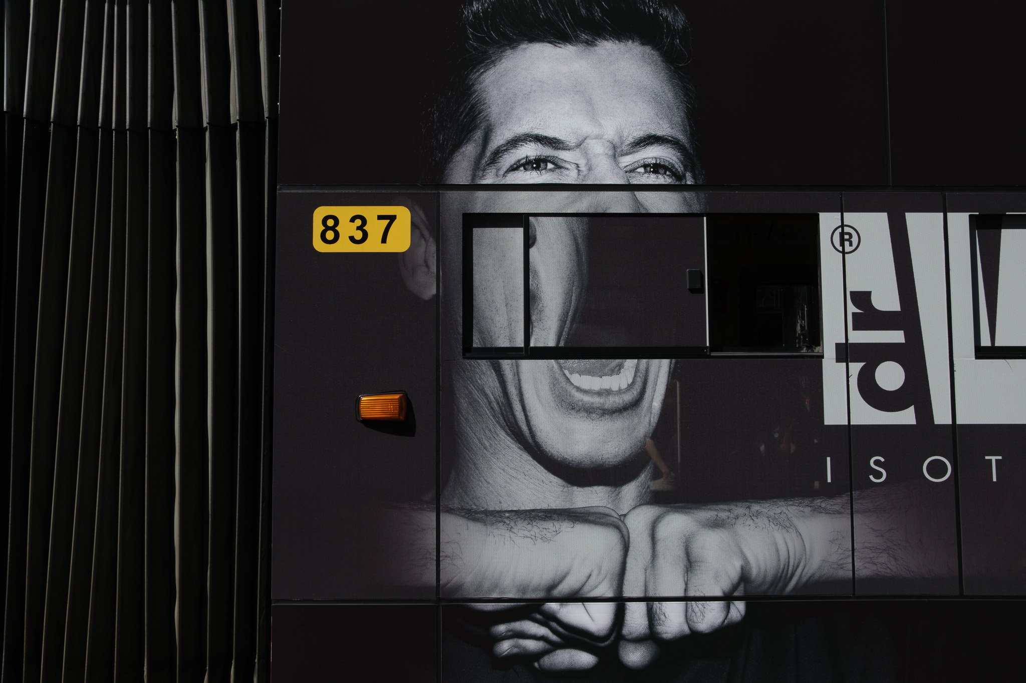 Black and white mural of a man's face with an angry expression and clenched fists, on the side of a building. The number 837 is displayed on a yellow background.