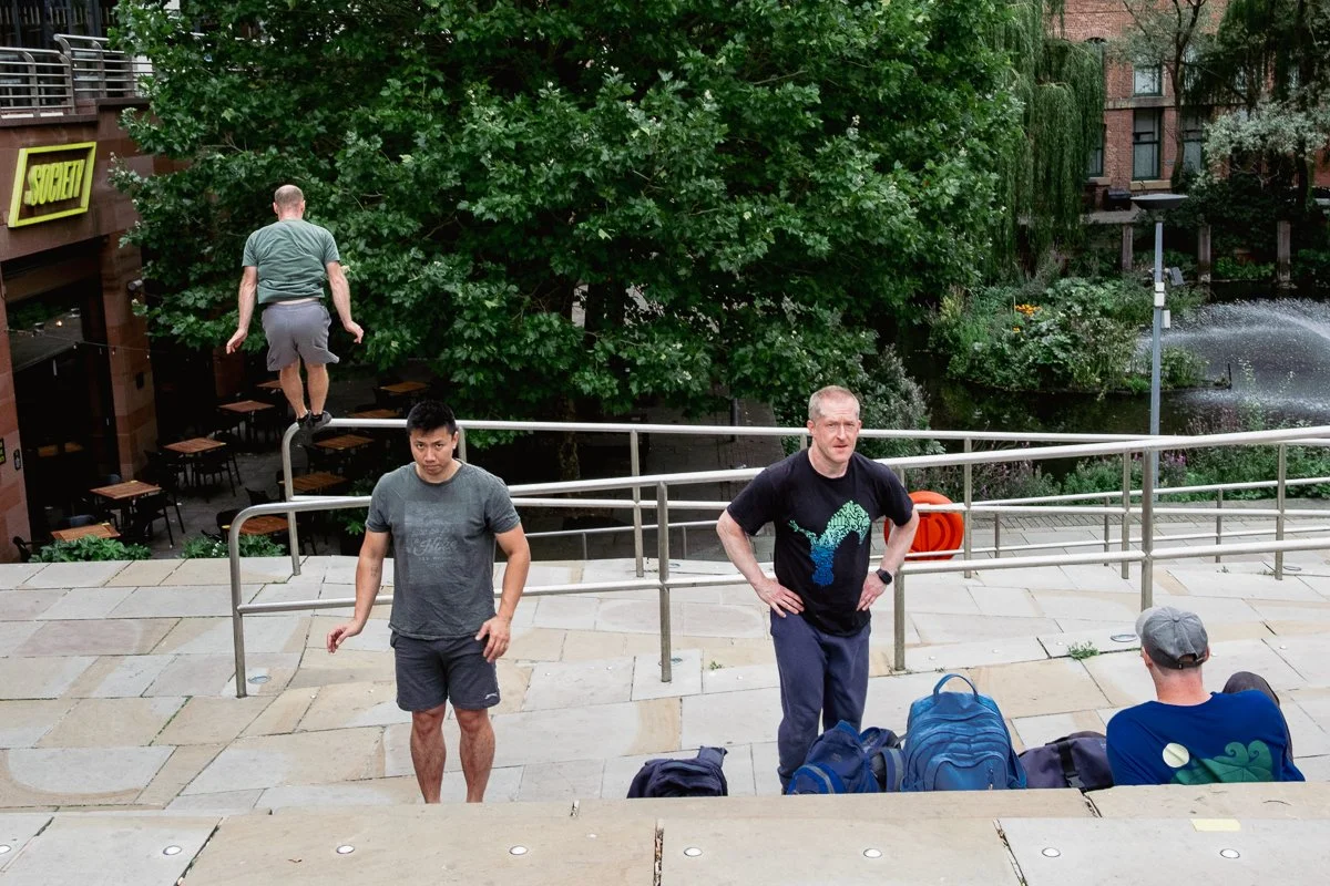 Four men are on an outdoor staircase with a metal railing. One man is sitting on the stairs, another is standing near the railing, a third is walking down the stairs, and the fourth is balancing on a handrail. There are backpacks on the stairs. In th