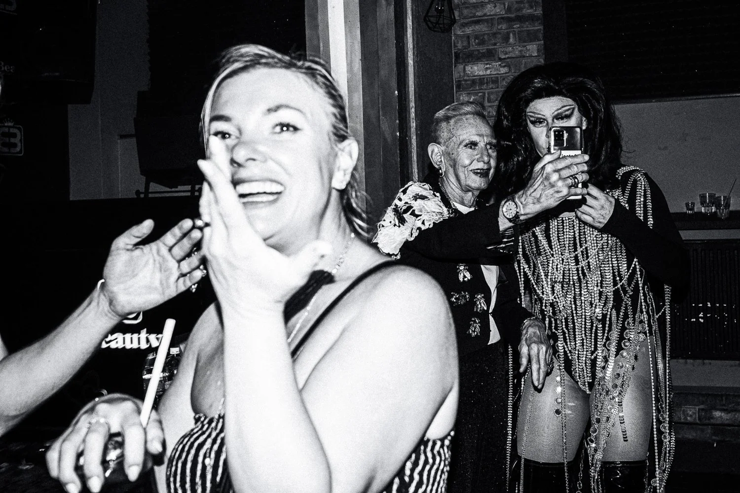 A black and white photo of three women in a lively social setting, with one woman at the forefront smiling and others in the background taking photos; the setting appears to be a bar or lounge with exposed brick walls.