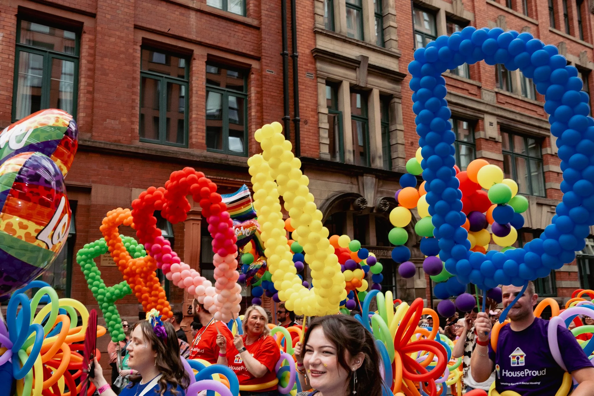 People participating in a pride parade with colorful balloon sculptures spelling out 'LOVE' against a red brick building.