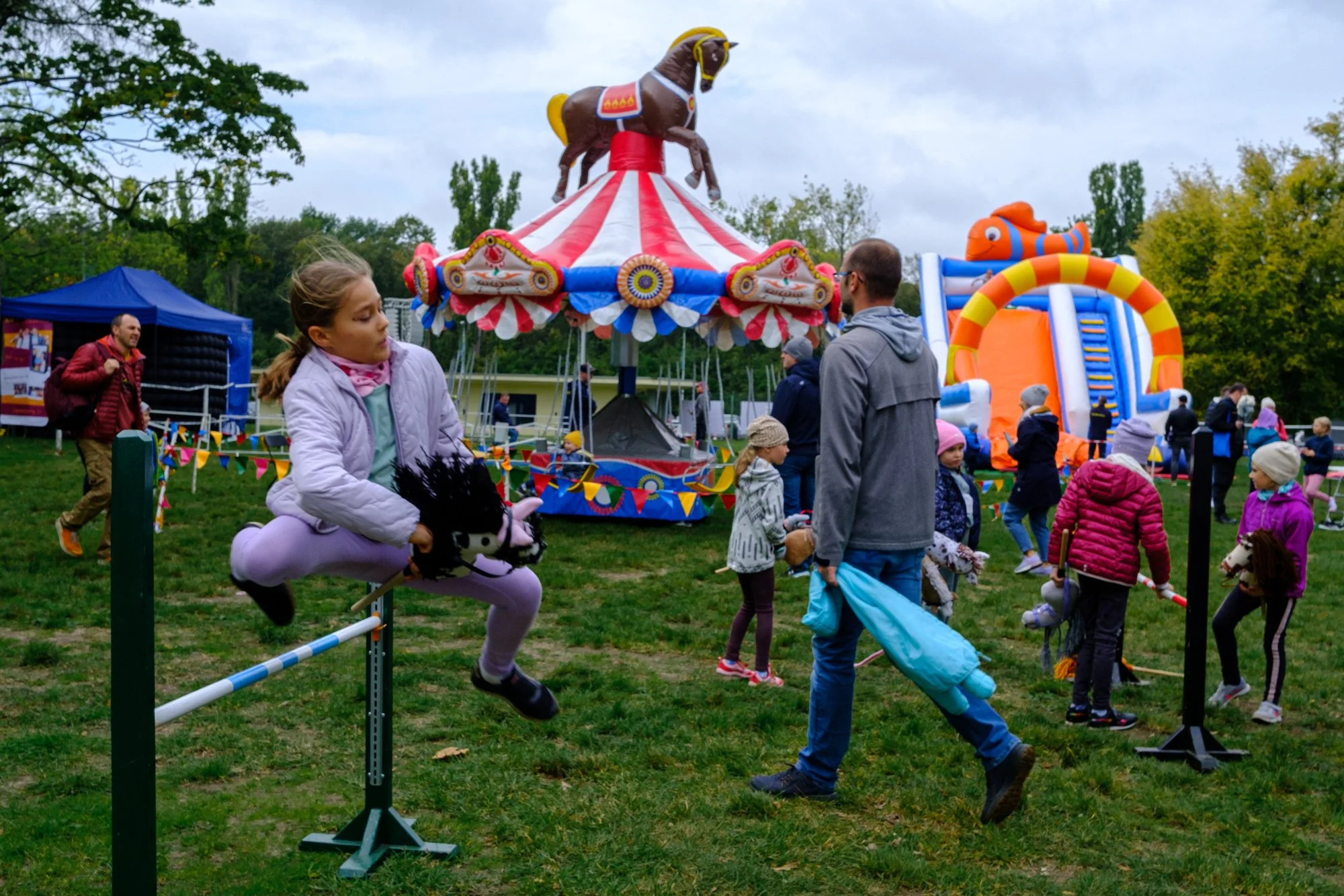 Children and adults enjoying a fairground with a carousel, inflatable slides, and games on a grassy field during cloudy weather.
