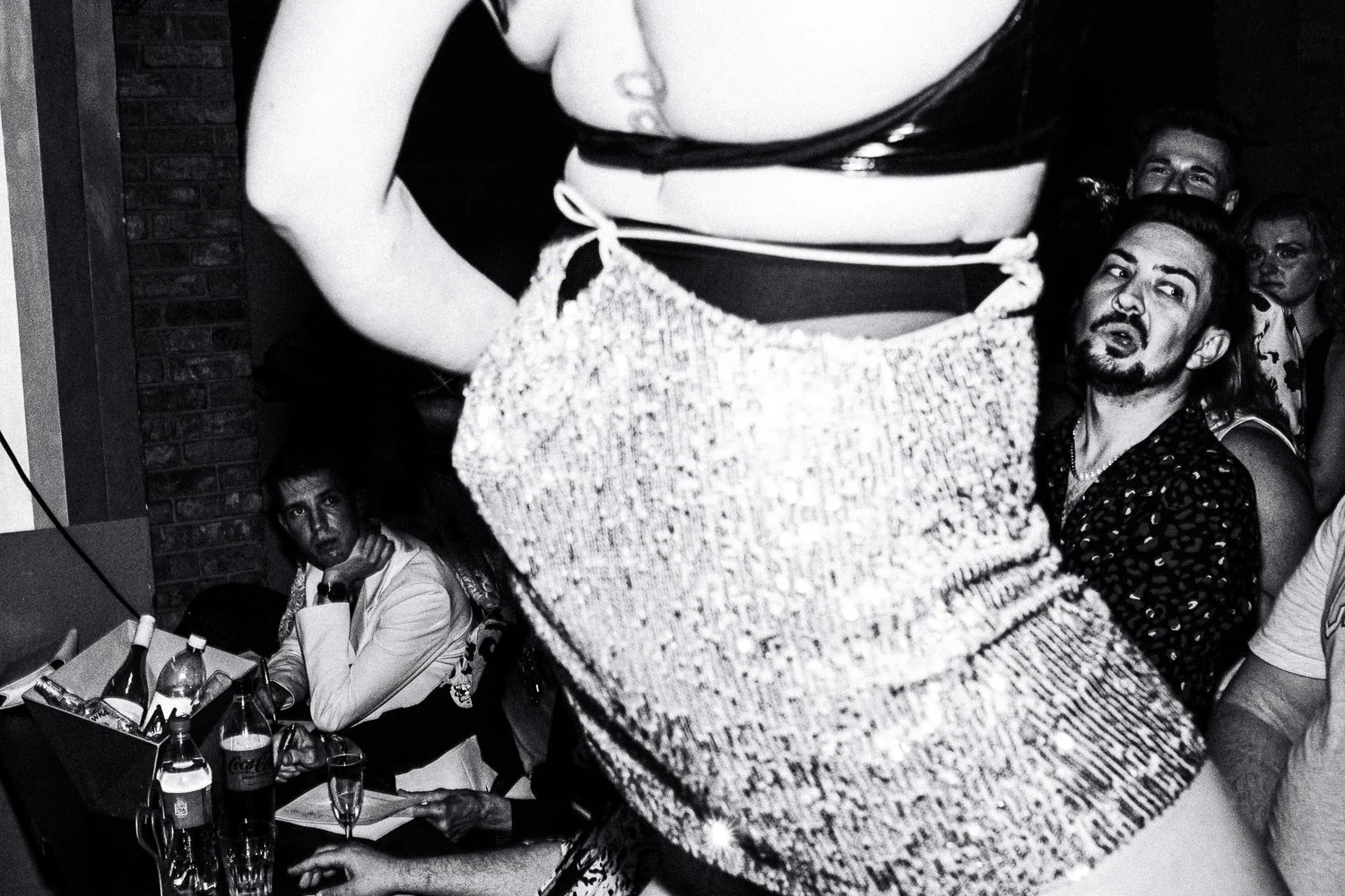 Crowd of people watching a performer on stage, with some individuals looking surprised or intrigued, sitting at a table with beverages.
