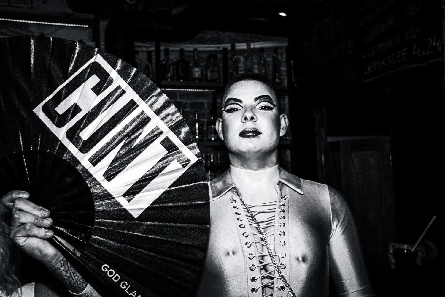 Person holding a fan with the words 'GAY PRIDE' written on it, in a bar or club setting, with bottles in the background, in black and white.