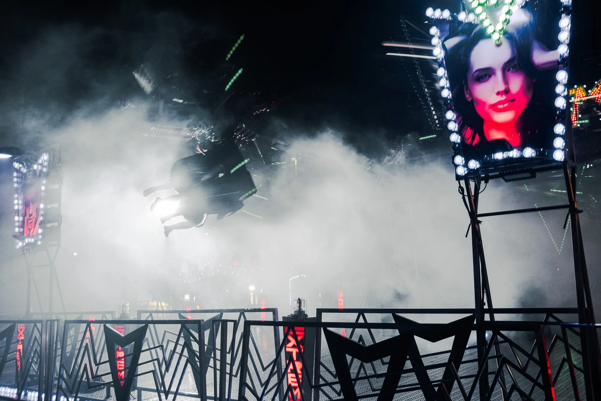 Amusement park ride with two people on a swinging chair suspended in the air, surrounded by mist or smoke, with colorful electronic screens displaying a woman's face with dark hair and bright makeup in the background.