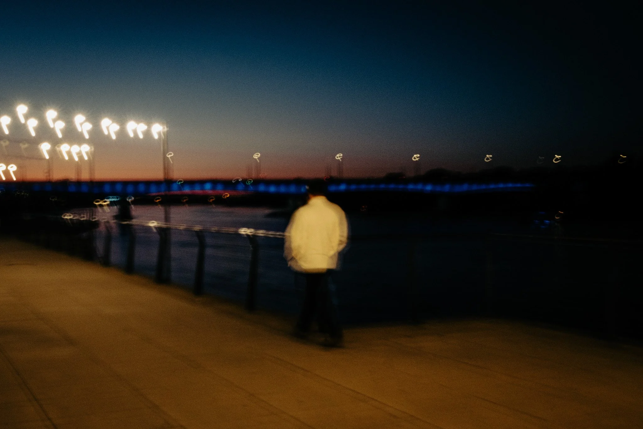 A person walking along a waterfront promenade at dusk or dawn, with a city bridge illuminated in blue lighting in the background, and blurred streetlights on the left.
