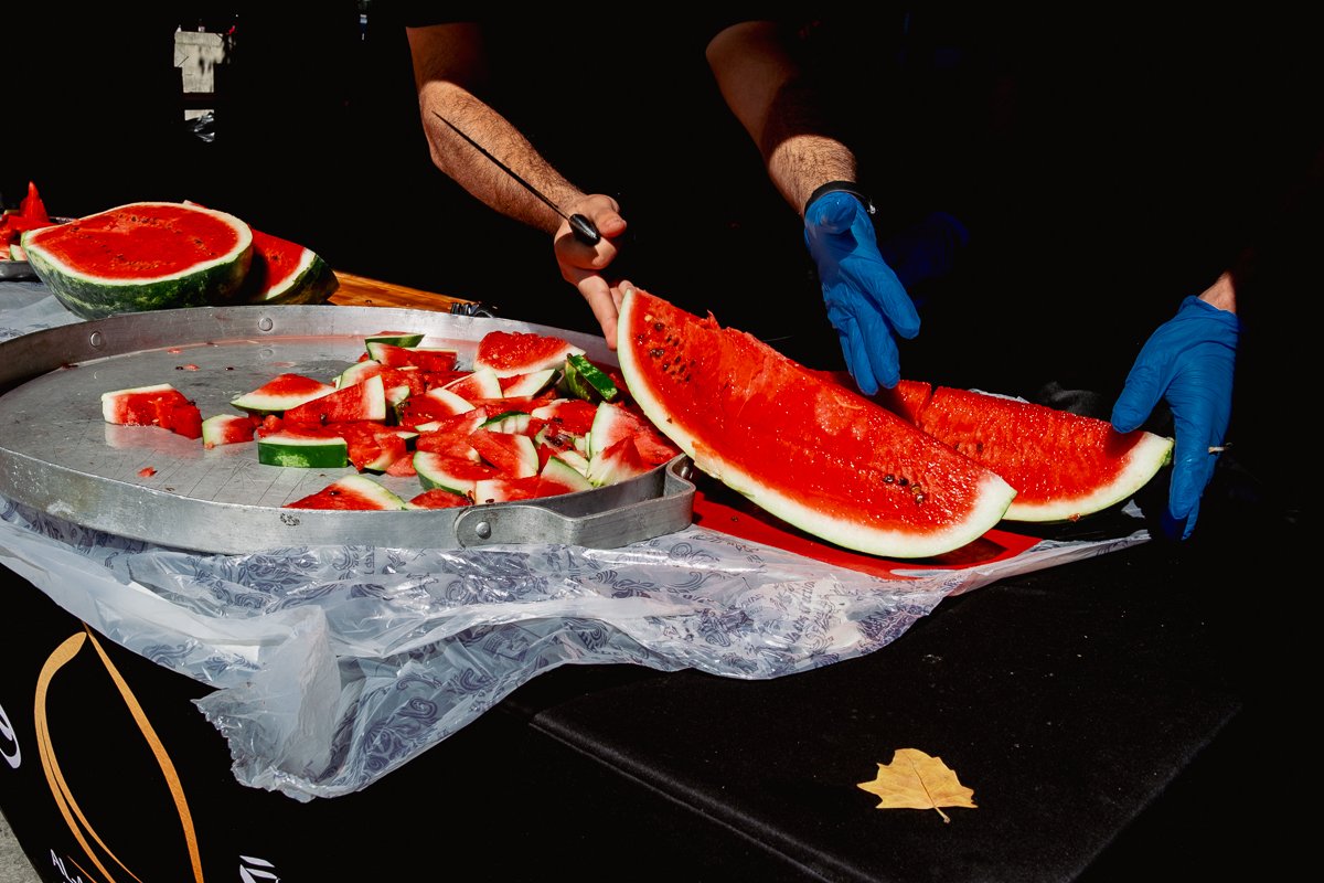 Person slicing a large watermelon with blue gloves while additional watermelon pieces sit on a tray, on a table covered with plastic, outdoors on a sunny day.