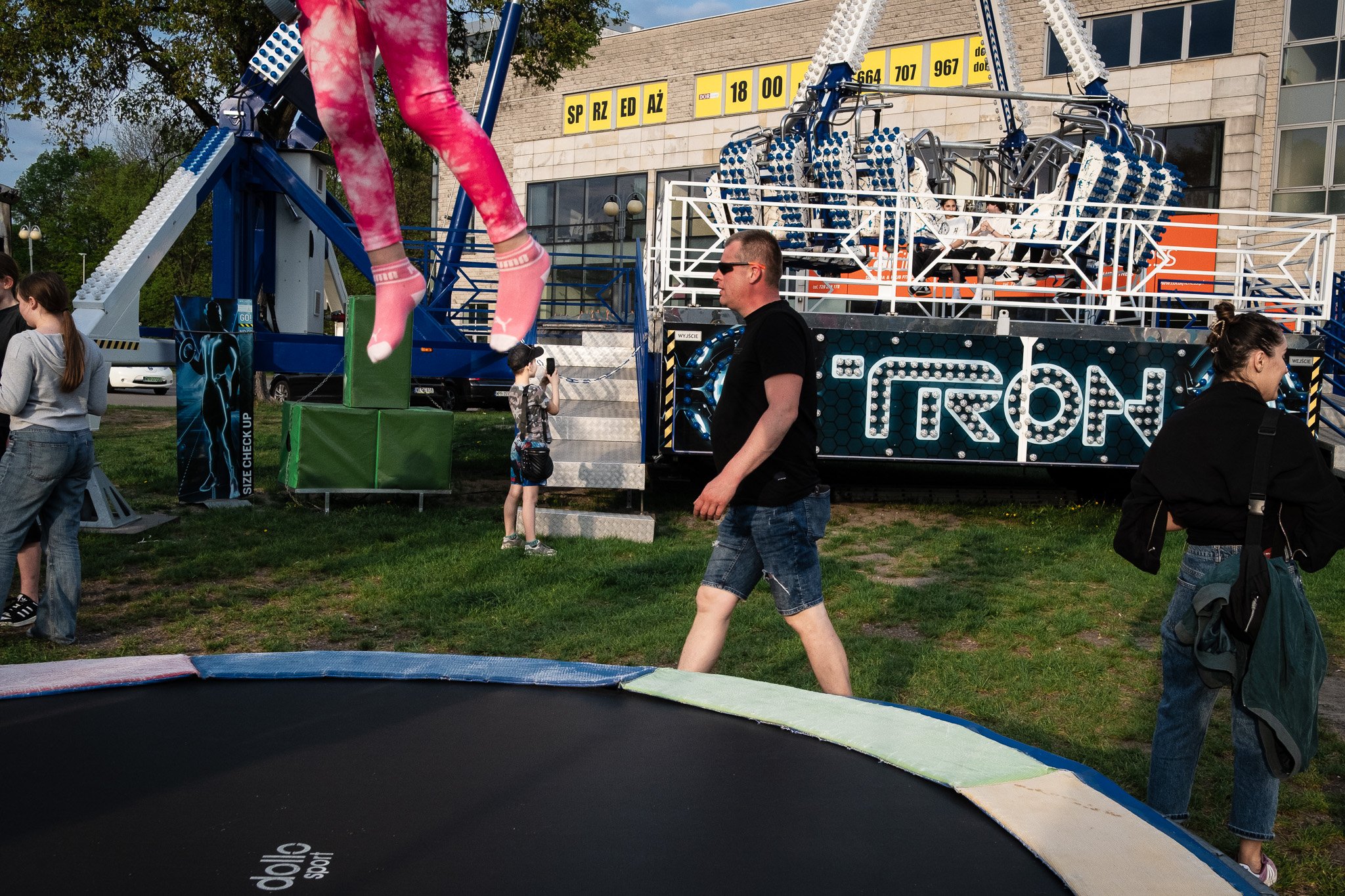 People at an amusement park or fairground with a blue and white ride in the background, a trampoline in the foreground, and a large poster of a girl in pink on the ride.