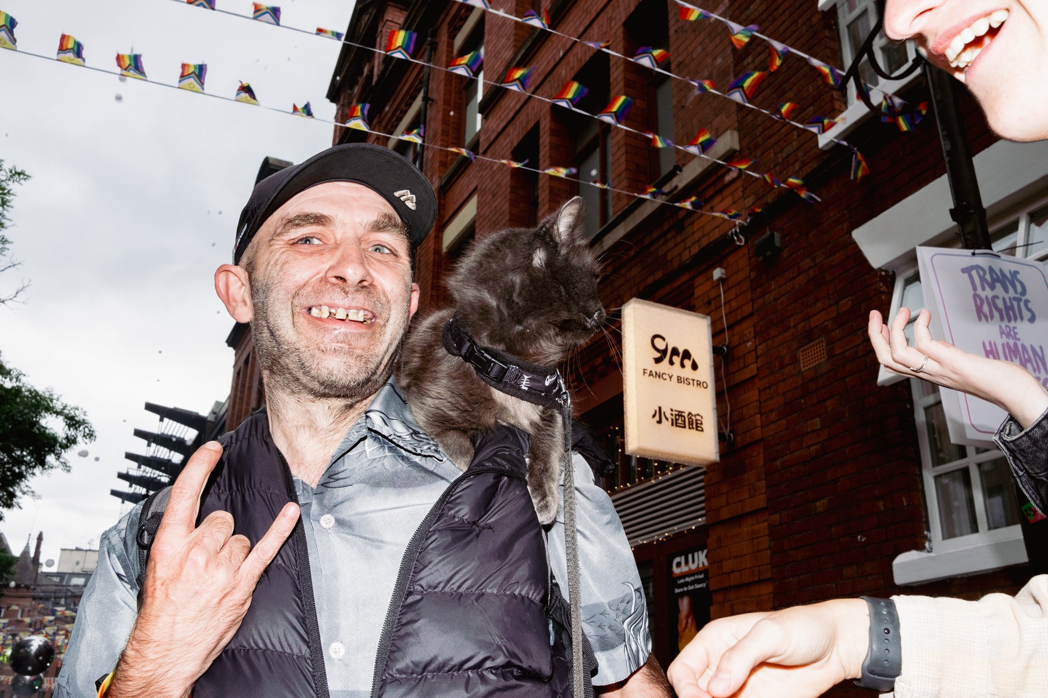 A man with gray hair, a beard, and sunglasses on his head, smiling and showing teeth, is holding a black cat on his shoulder. The man is making a hand gesture with his fingers. Outside, with a brick building in the background, colorful pride flags ha