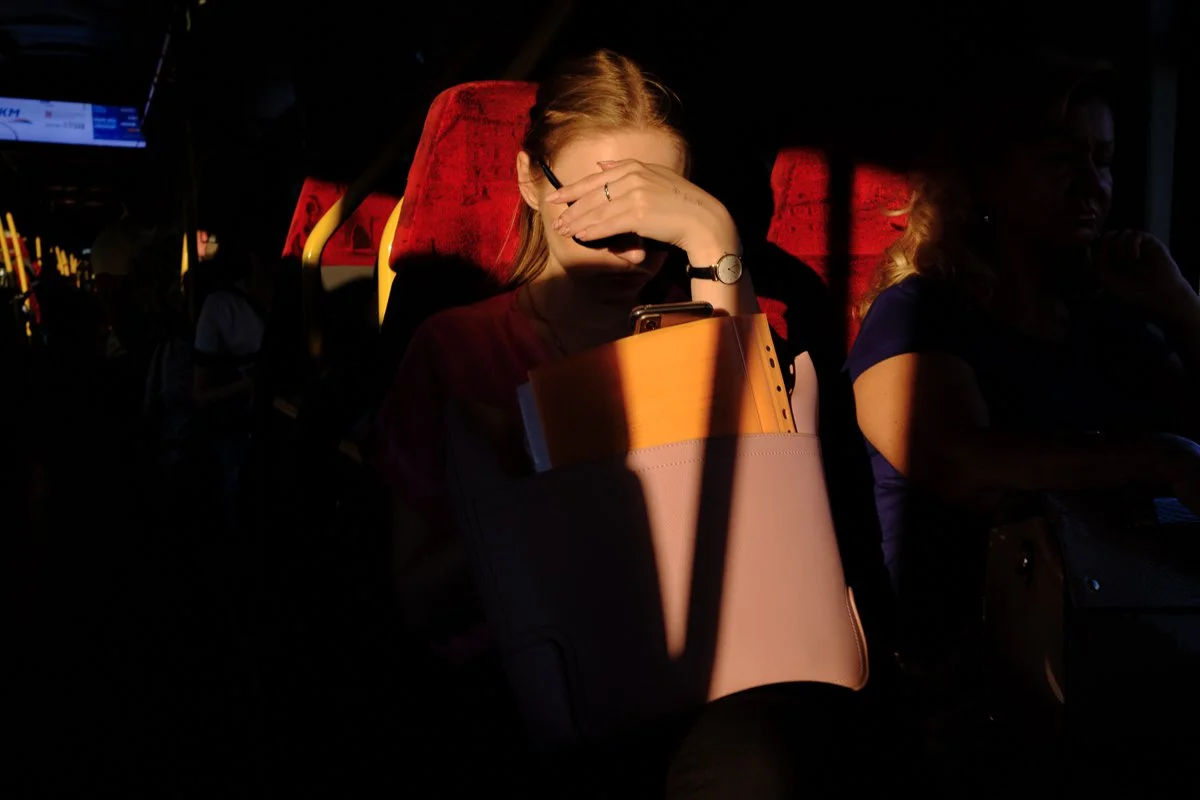 A woman sitting on a bus, using her phone with the sunlight casting a shadow and highlighting her face and hand.