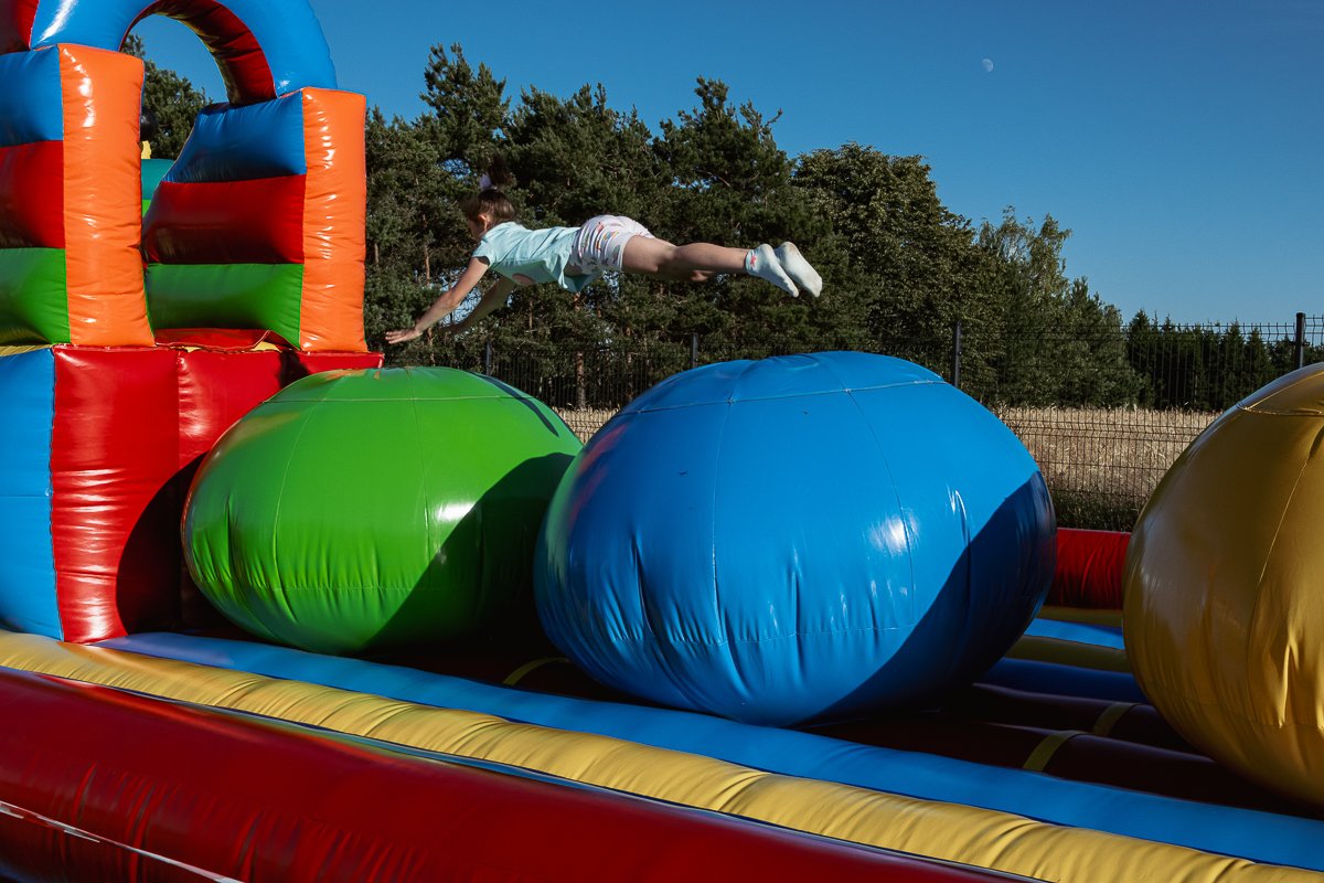 A girl is sliding or leaping across large inflatable balls on an outdoor inflatable obstacle course, with trees and a fence in the background and clear blue sky above.