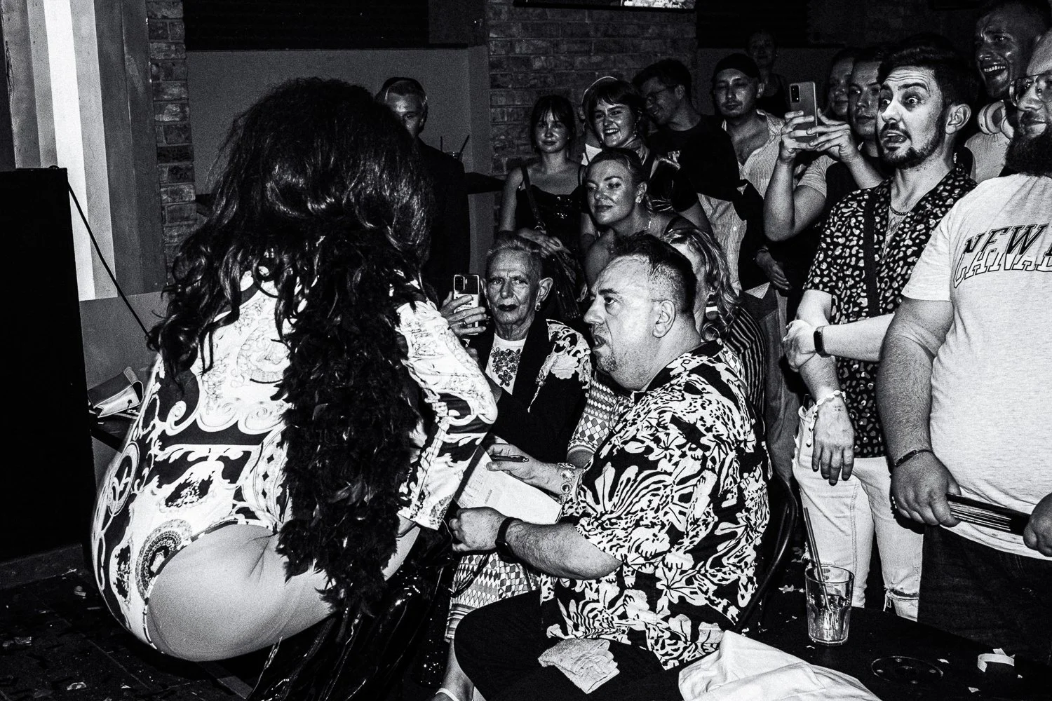 A black-and-white photo showing a woman in a patterned dress leaning over a man seated at a table, who appears to be yelling or arguing. The scene is crowded with people, some taking pictures and others watching, in a social setting or event, with a 