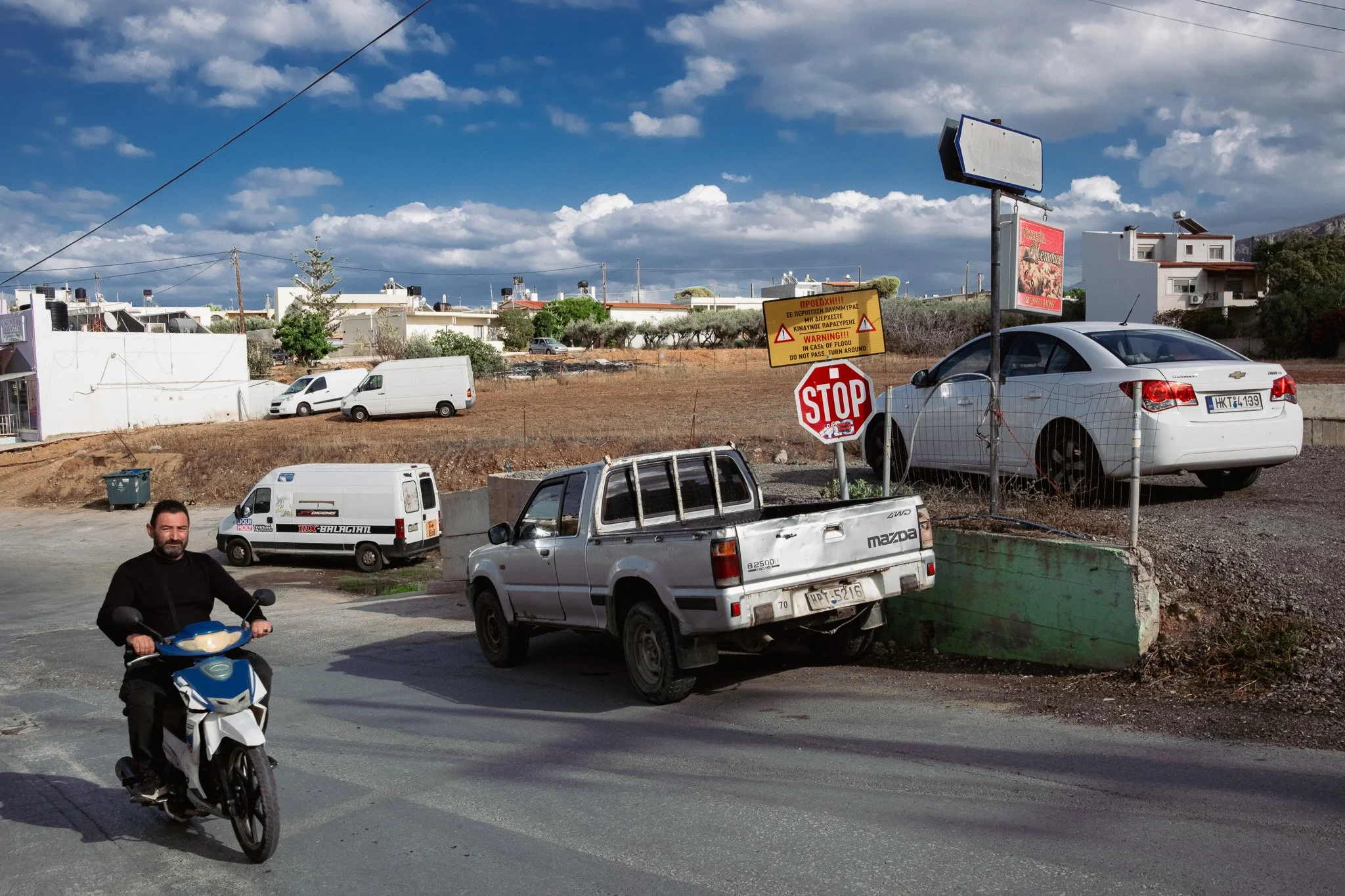 A man riding a scooter on a street with parking areas and vehicles, including a white pickup truck, white cars, and vans. There are multiple signs, including a STOP sign and warning signs, near a fenced area under a partly cloudy sky.