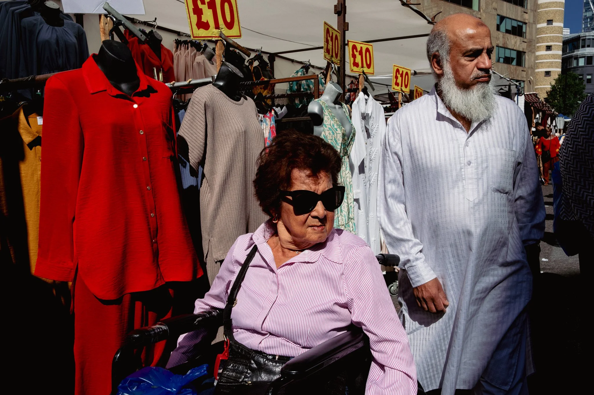 An elderly woman in a wheelchair wearing sunglasses and a pink striped shirt stands beside an elderly man with a white beard wearing a white traditional outfit at an outdoor clothing market. Behind them, there are racks of clothes with red and yellow