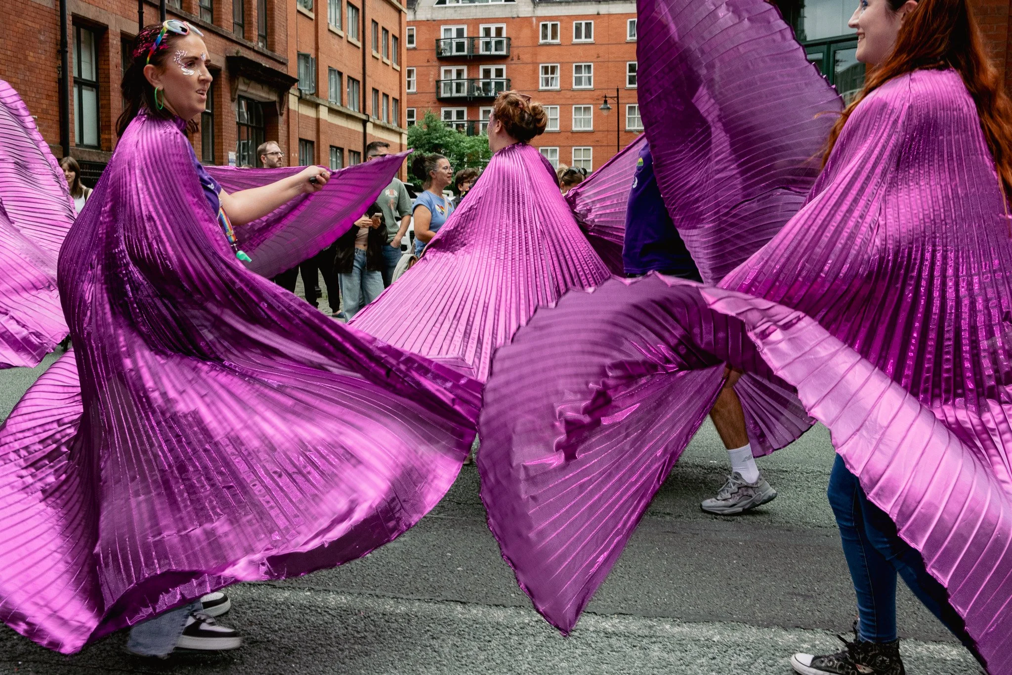 People participating in a street parade or gathering, holding large purple fabric pieces, with a brick building in the background.