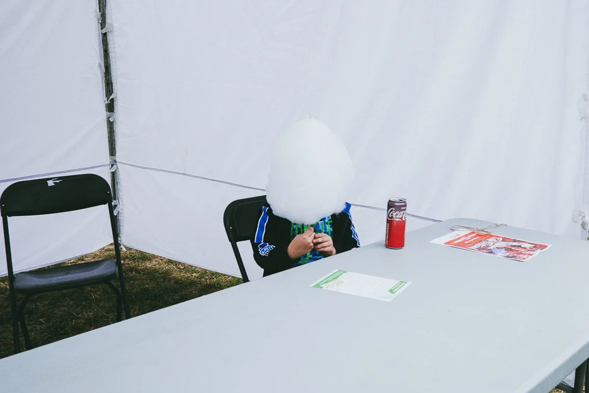 Person sitting at a table in a white tent with a large cloud of cotton candy covering their face, a can of Coca-Cola, and some papers.