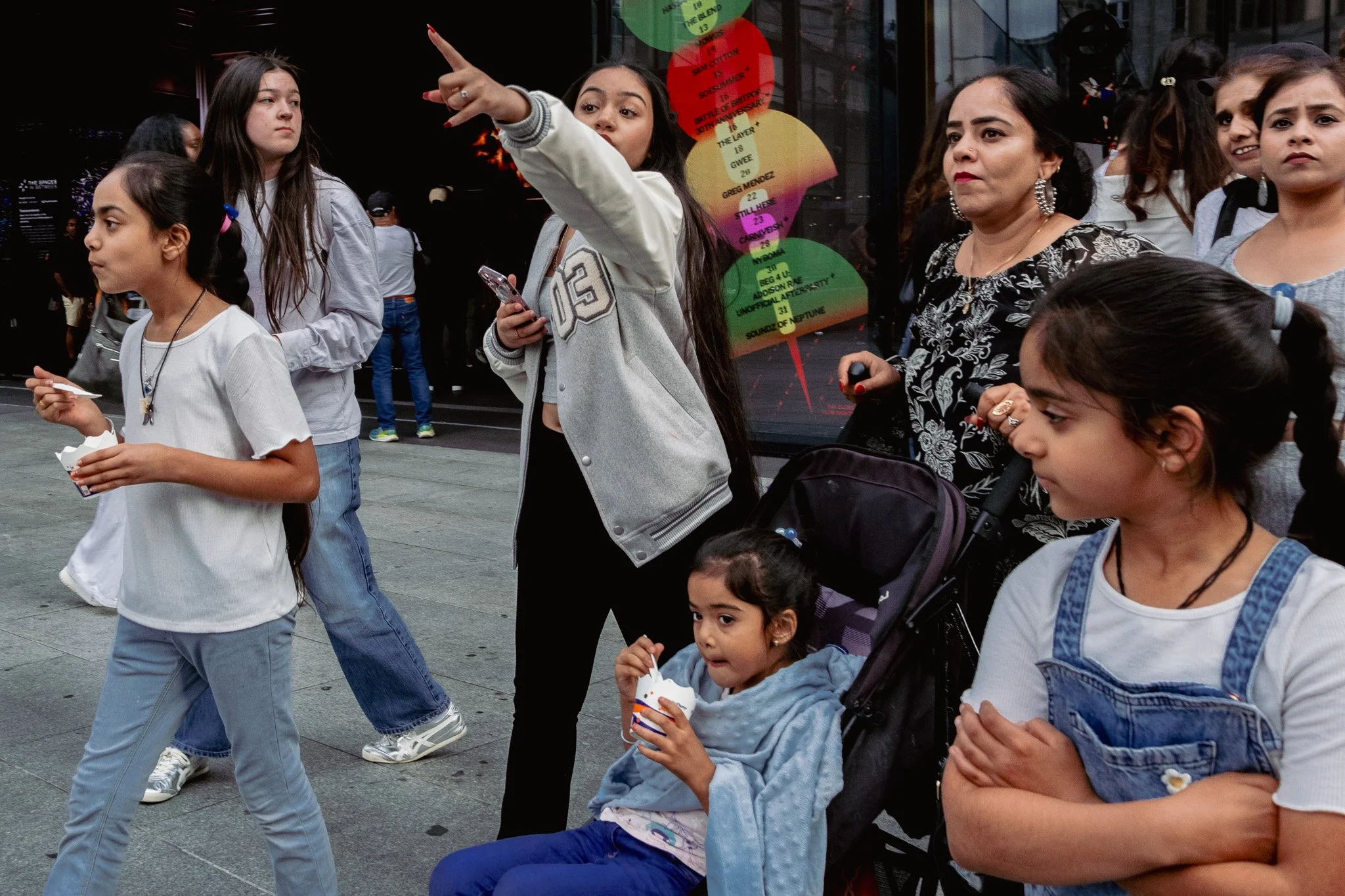 Group of children and women standing on a city street, some eating food from small containers, one girl in a stroller, and a woman pointing with her finger towards something off-camera.