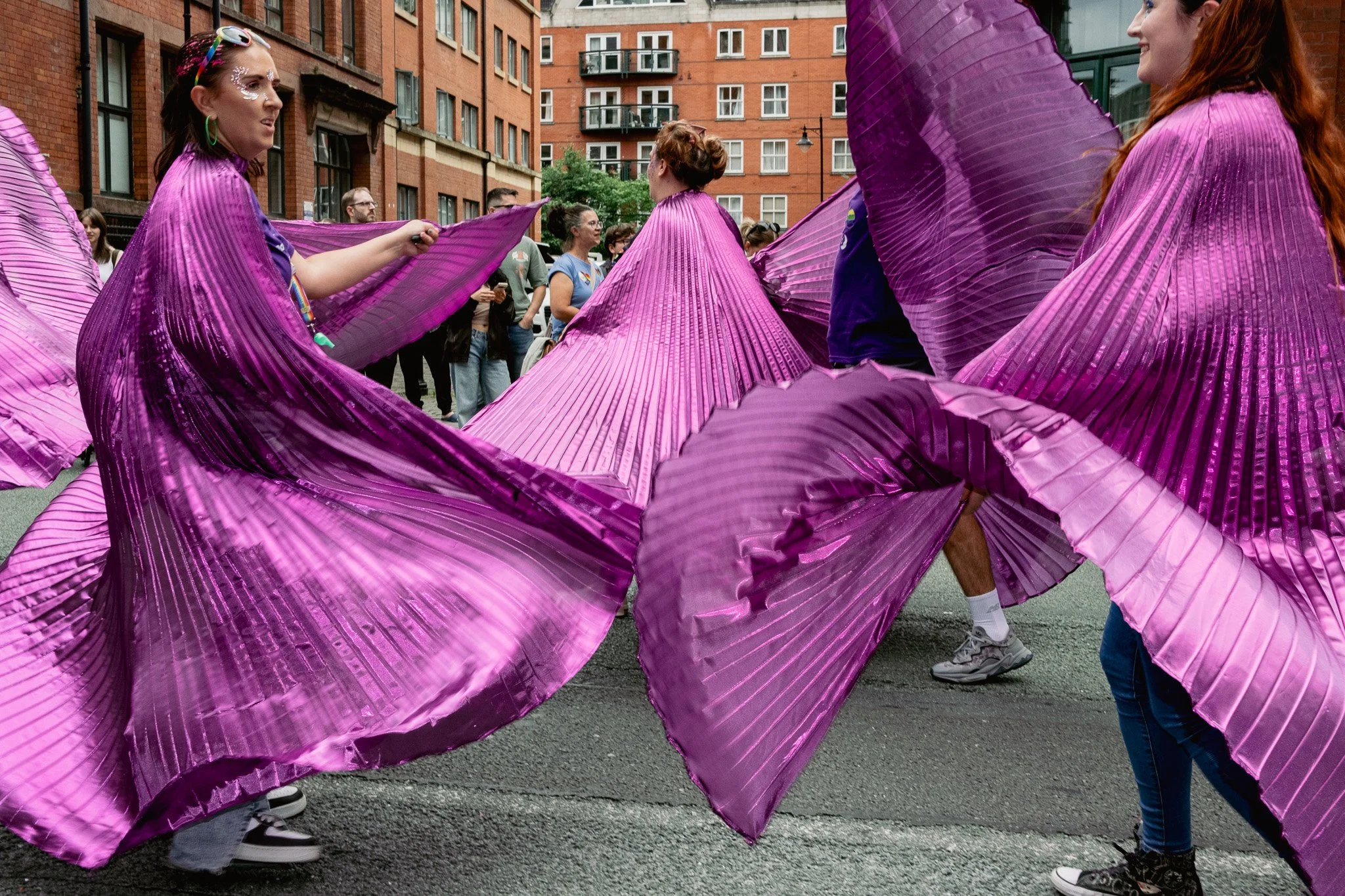People dancing in colorful metallic purple costumes with large pleated skirts at an outdoor event, with a brick apartment building in the background.