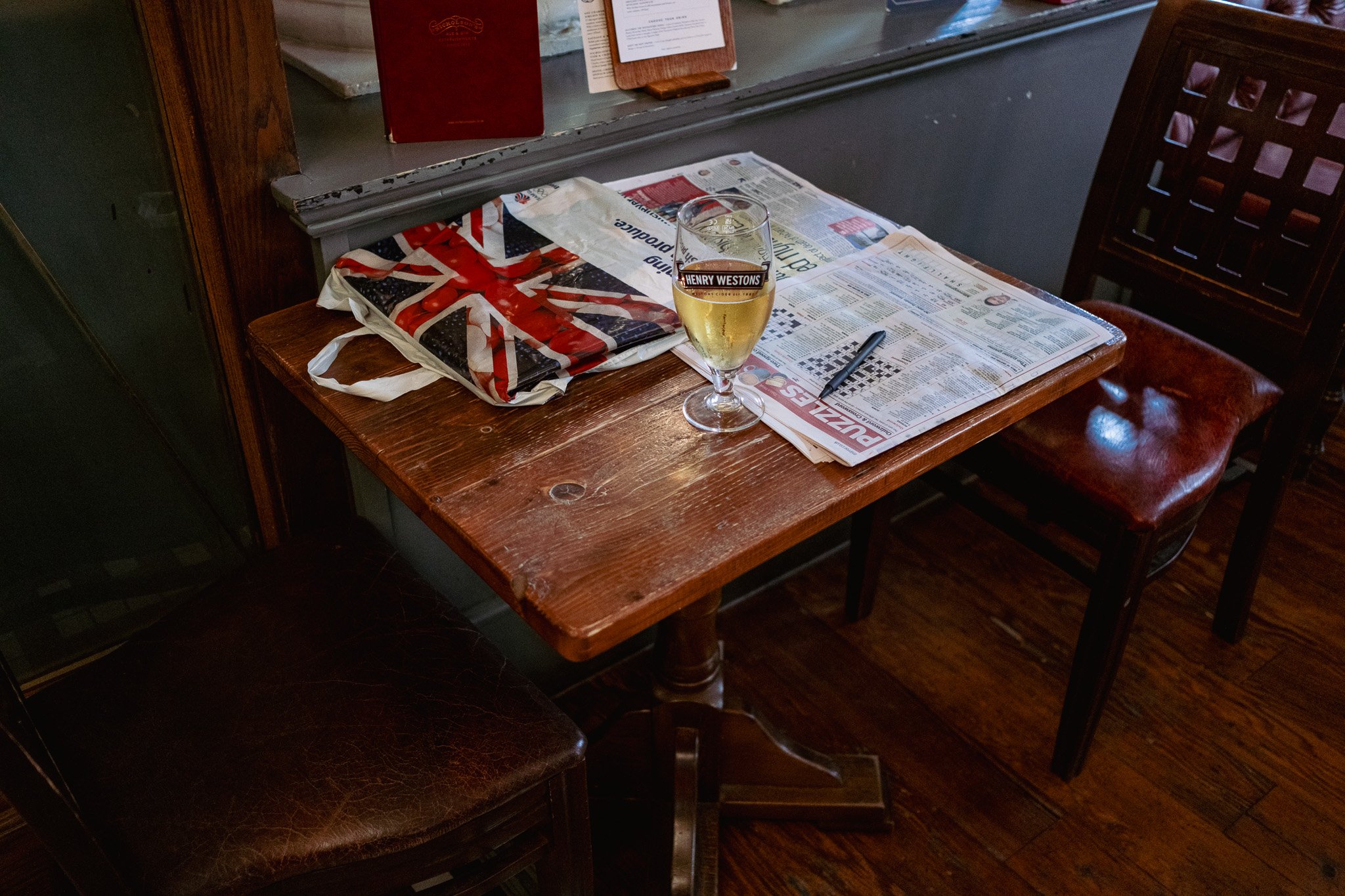 A wooden table with a British flag-themed bag, a glass of beer labeled Henry Westons, a newspaper, and a pen. There are two chairs, one with a leather seat.
