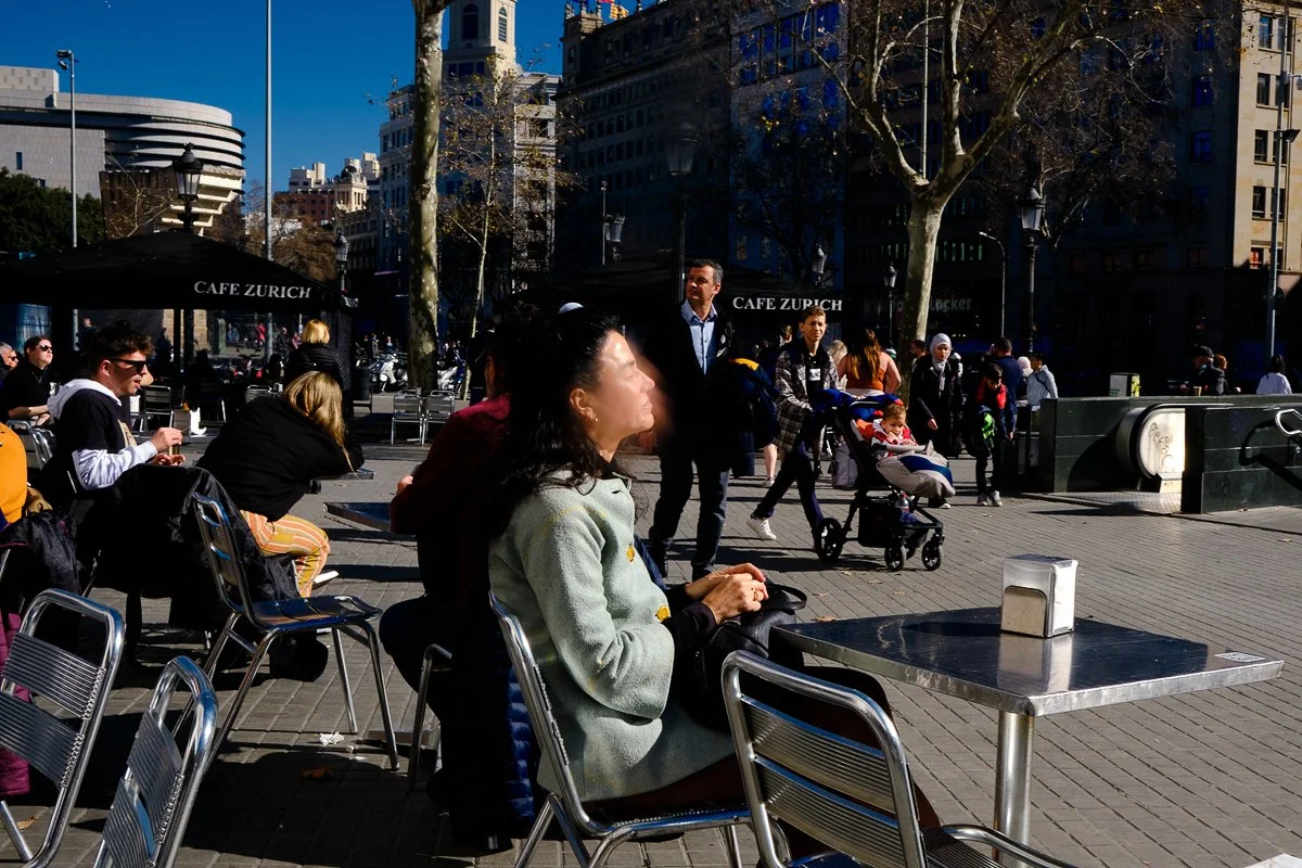 People sitting and walking at outdoor Cafe Zurich in a city plaza during daytime with tall buildings and leafless trees.