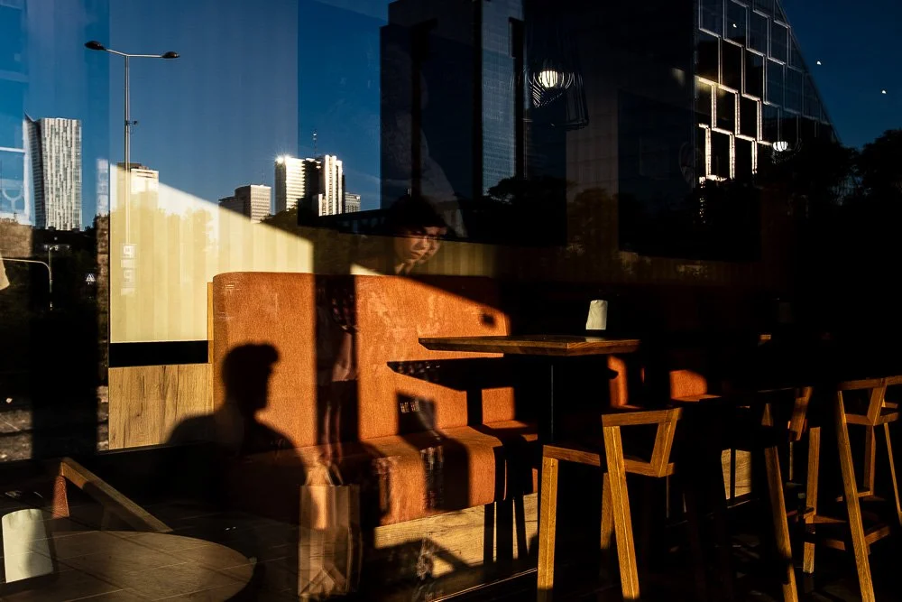 Reflections of city buildings and a person sitting inside a cafe at dusk, where the interior is visible with wooden chairs and tables.