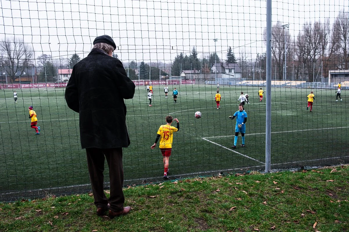A man watching children play soccer from behind a fence on a sports field, with children wearing colorful uniforms and a goalkeeper in blue.