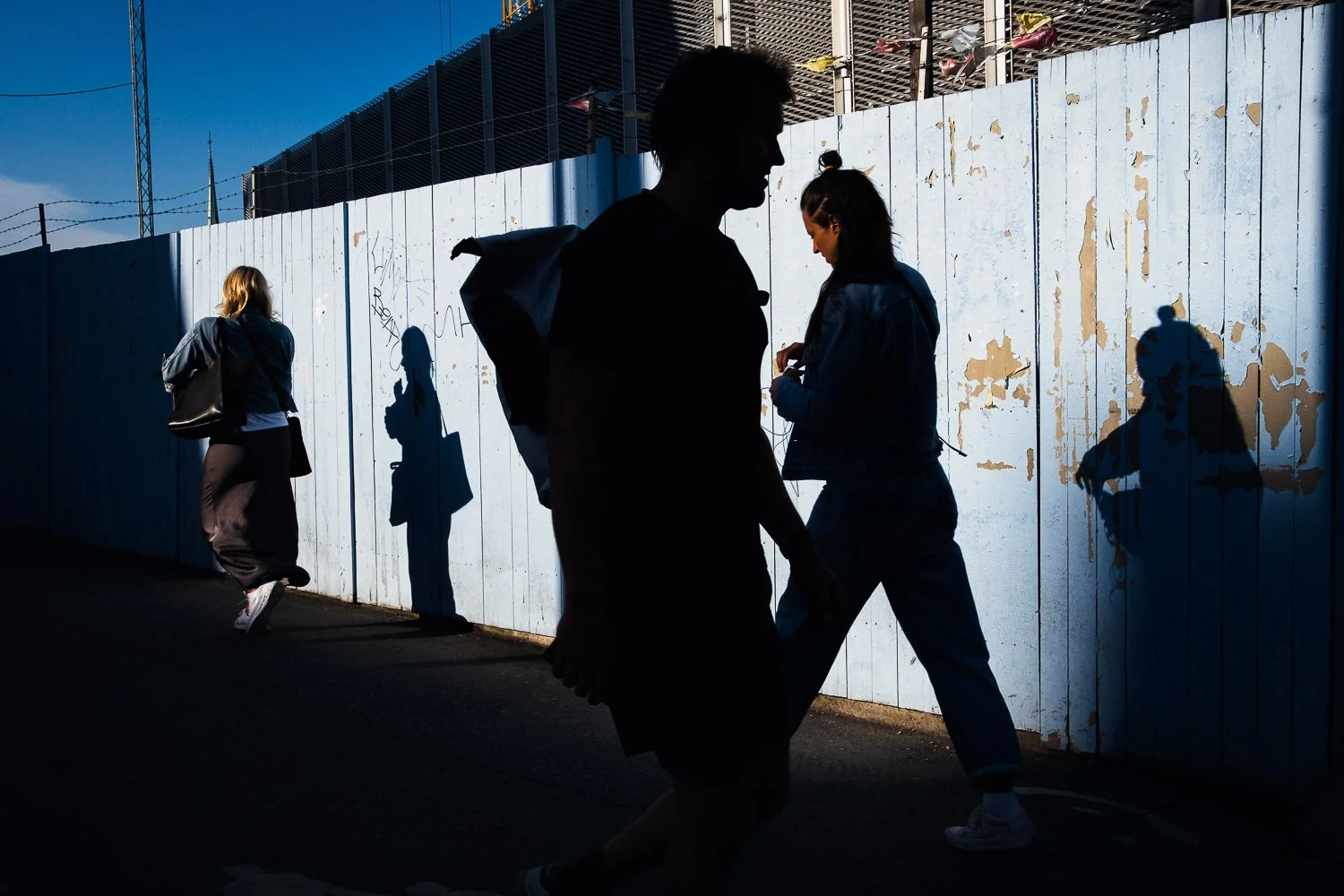 People walking past a wooden fence with their shadows cast on it during daytime. The silhouettes show the contours of the individuals, including a man and women, with a cityscape and blue sky in the background.