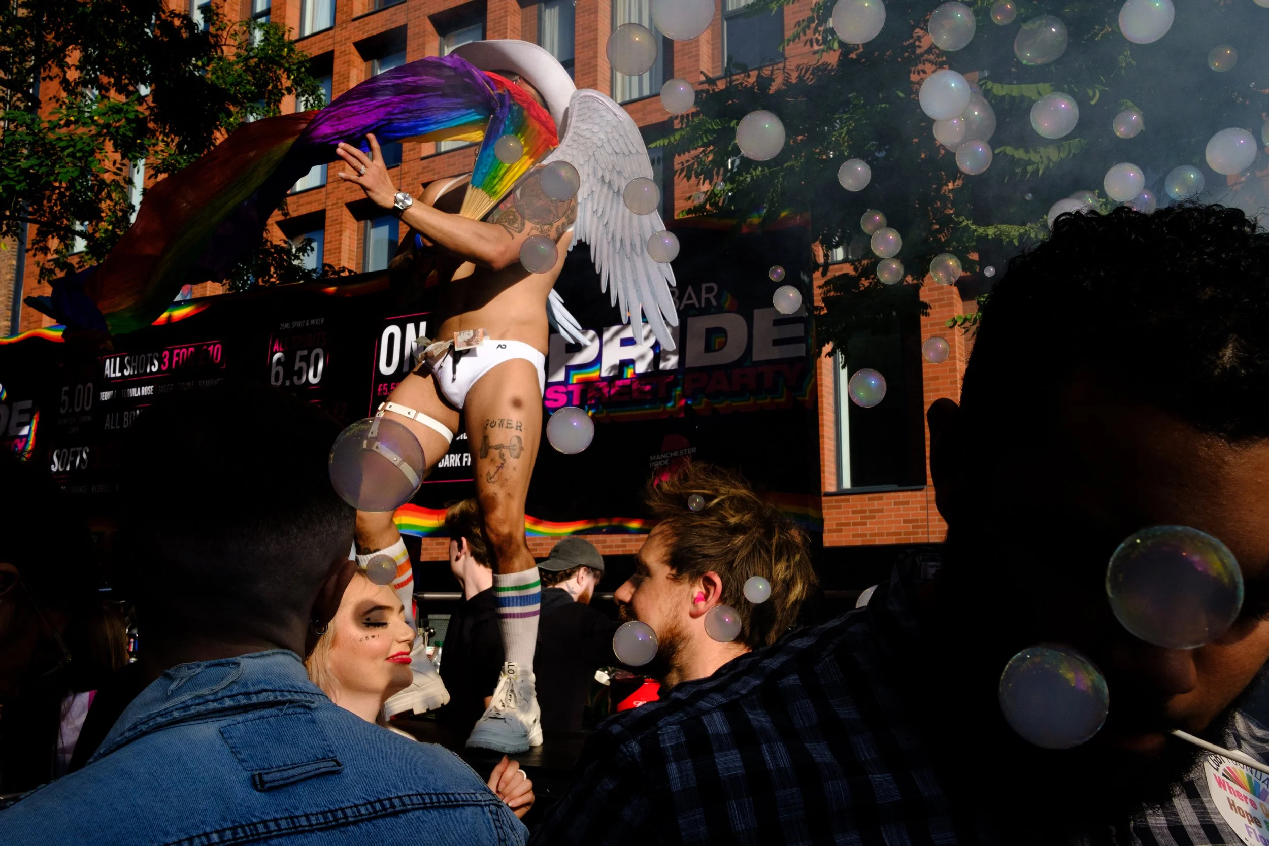 A person with angel wings and rainbow rainbow-colored rainbow mask performing on a float during a pride parade, with spectators watching, balloons floating around, and a brick building in the background.
