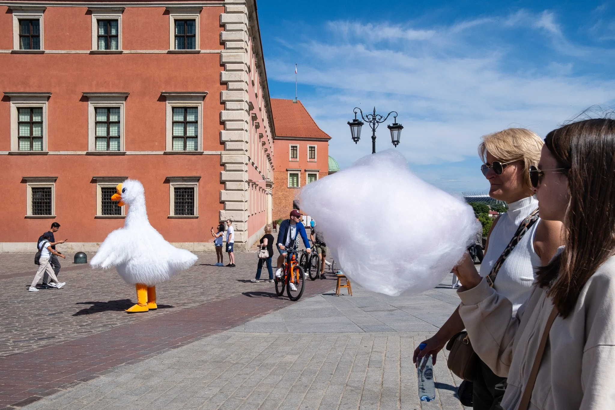 People on a city street with a person dressed in a duck costume and a woman holding a large piece of cotton candy.
