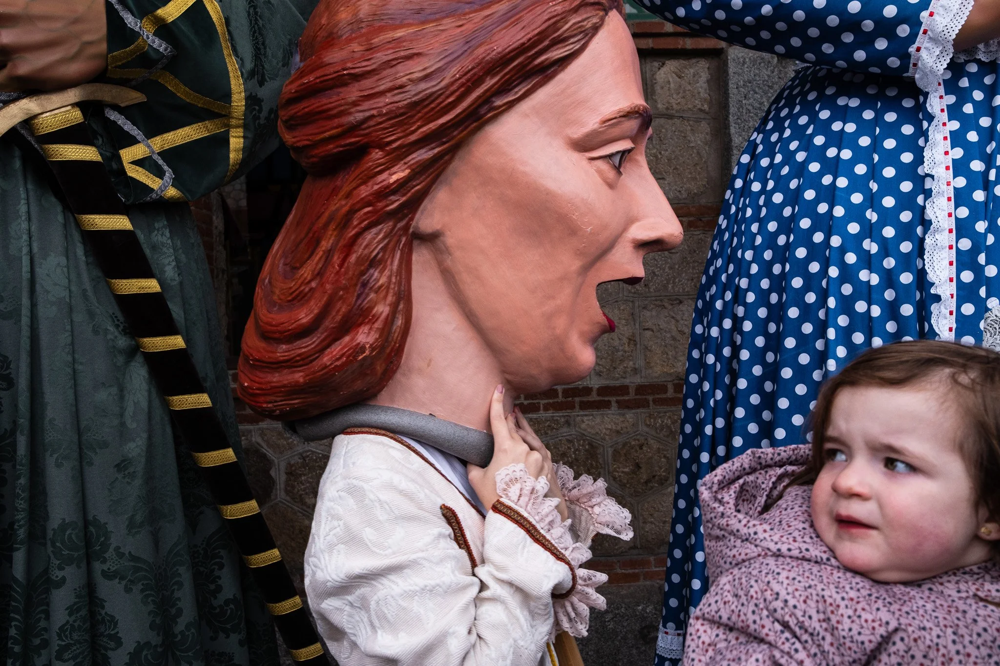 A large face mask resembling a woman with styled red hair and red lipstick, surrounded by people, including a child, in front of a brick wall.