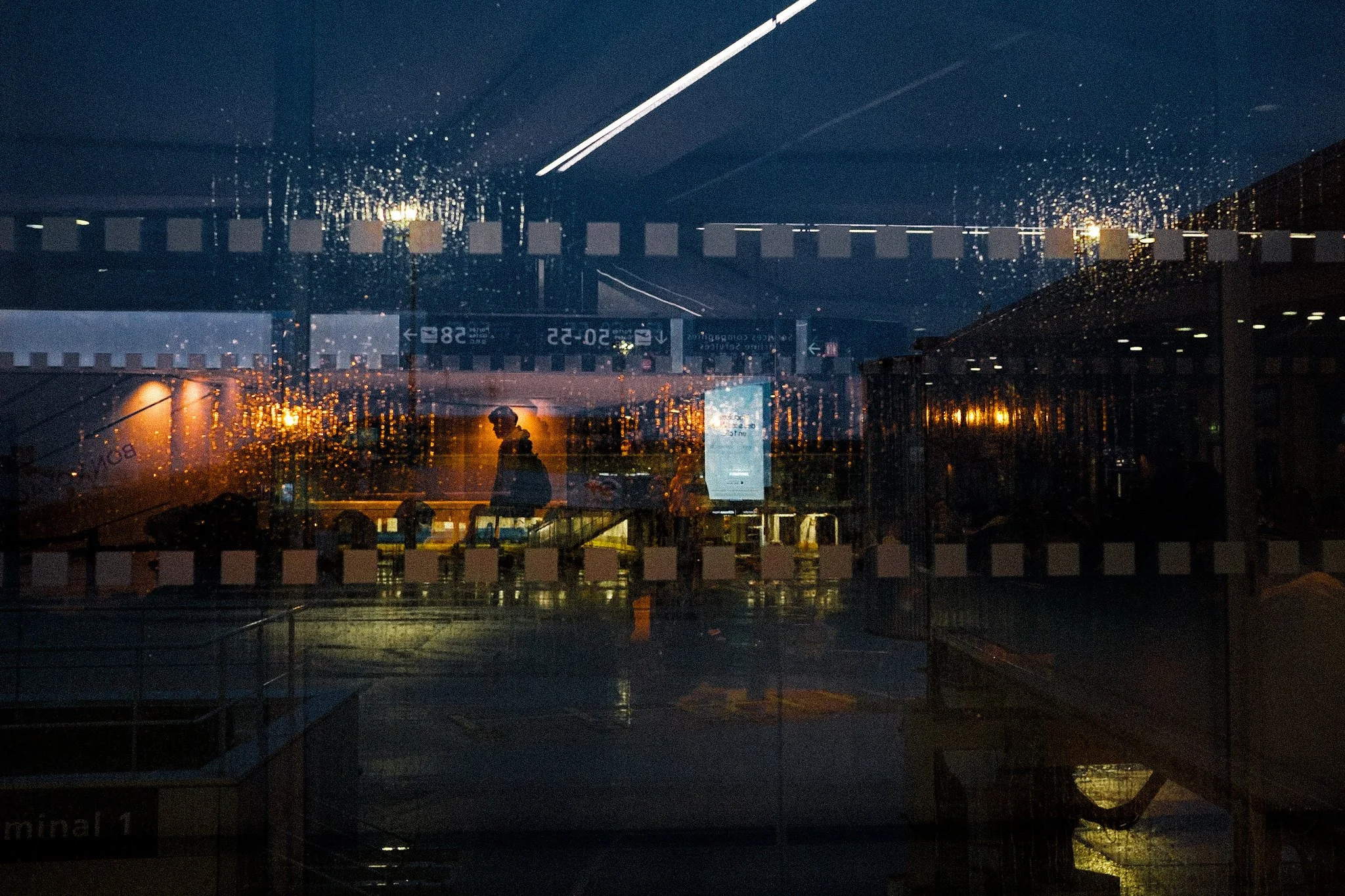 People walking through an airport terminal at night, visible through a rain-covered glass window with reflections of interior lights.