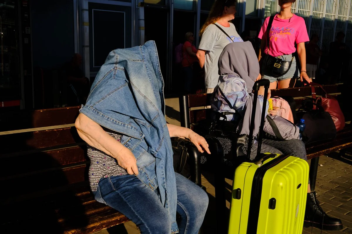 A woman with a denim jacket draped over her head sitting on a bench at the train stain, surrounded by luggage and bags, with two young women standing nearby.