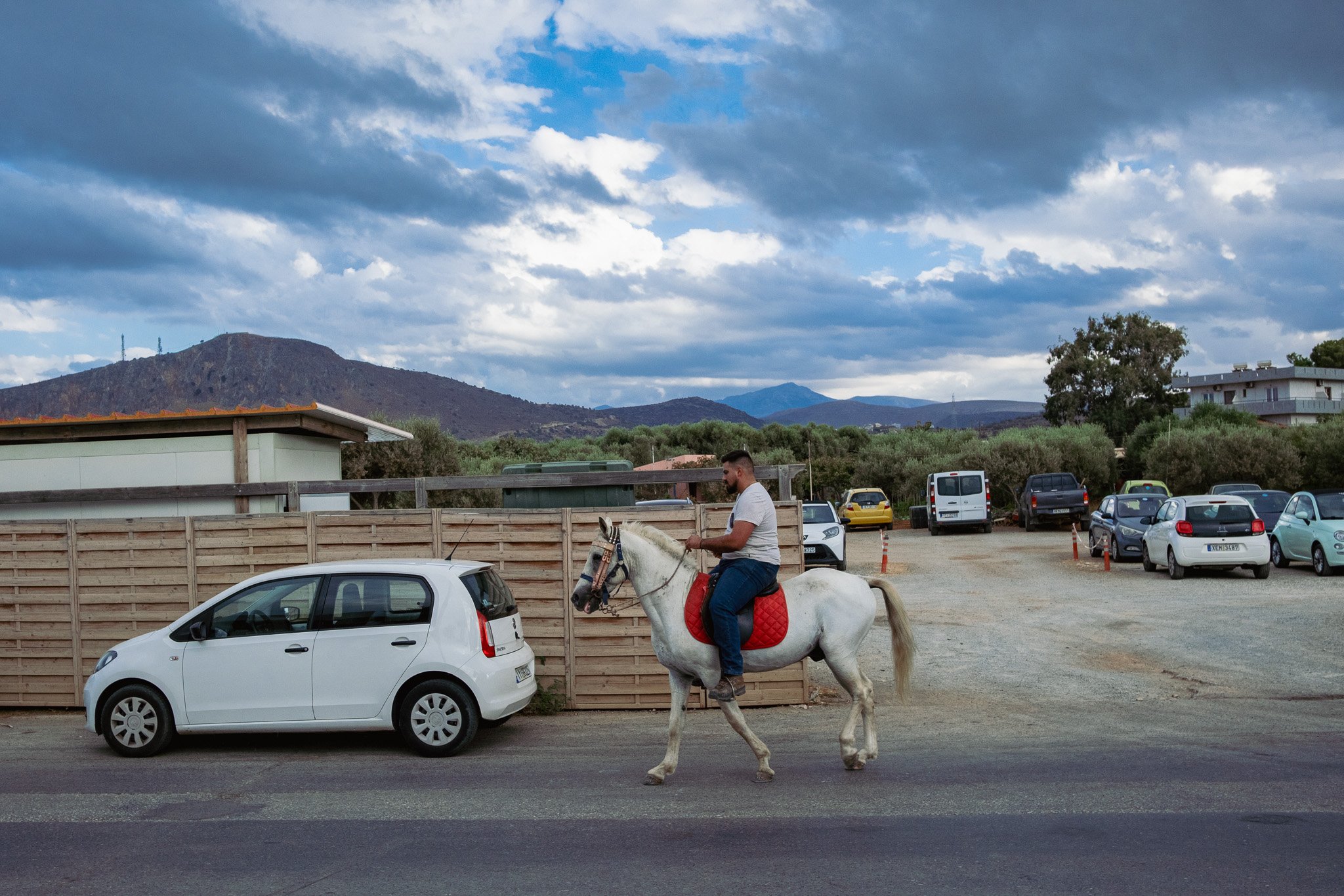 A man riding a white horse with a red saddle and bridle past parked cars and a wooden fence, with mountains and a cloudy sky in the background.